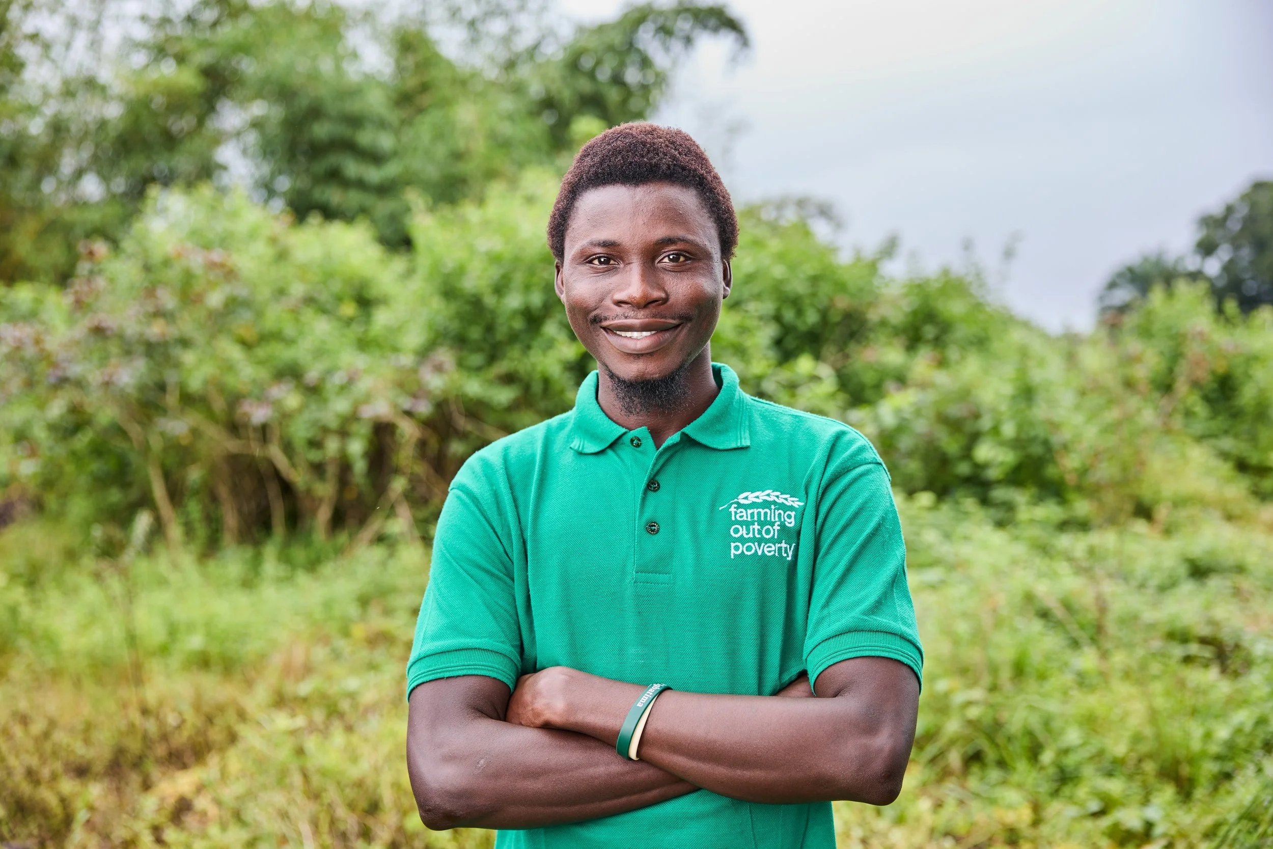 Smiling young man with arms crossed, wearing a green polo shirt with the logo 'farming out of poverty,' standing outdoors in a green, bushy area.