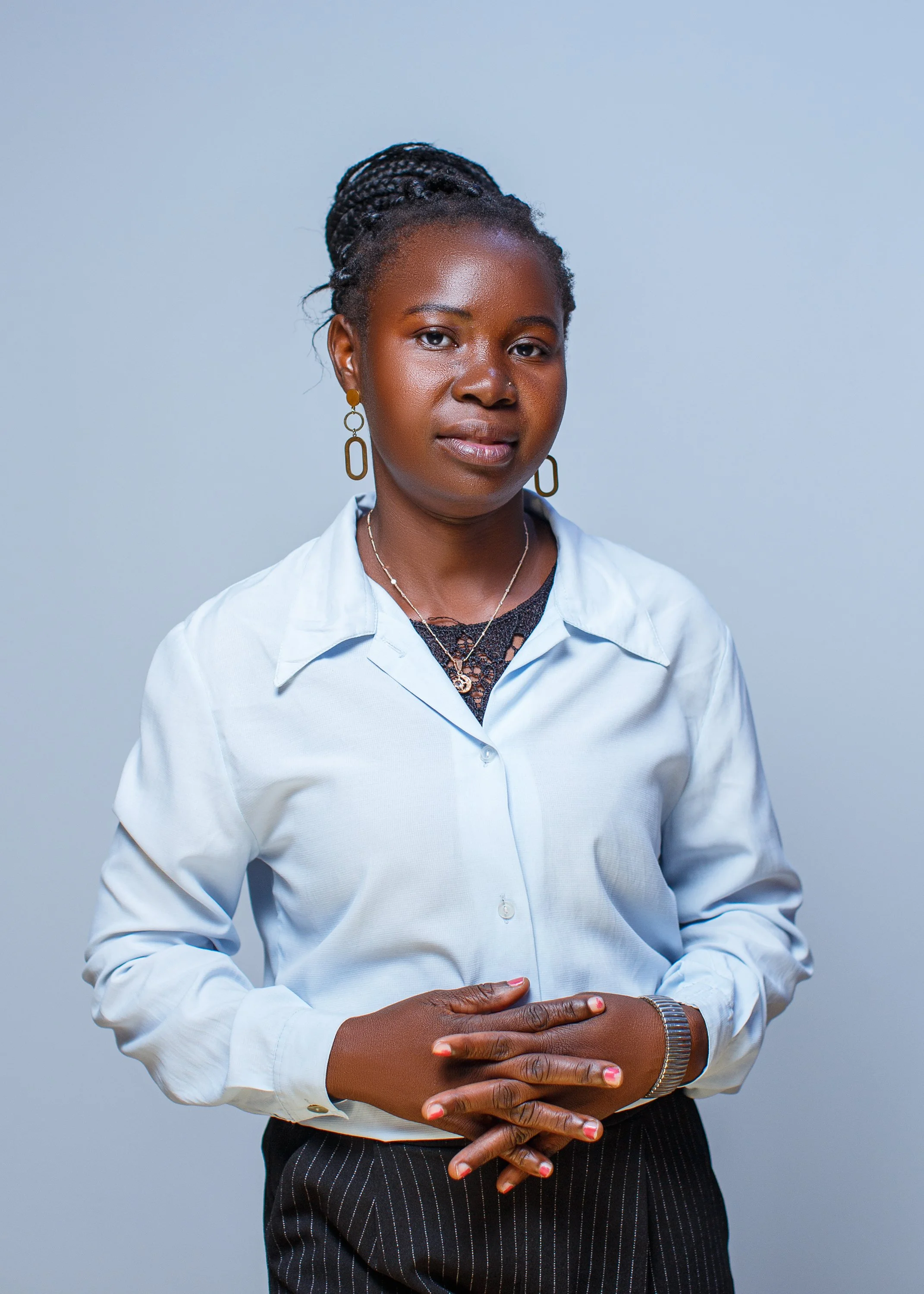 A woman with braided hair wearing a white shirt, black pinstripe pants, and jewelry, standing against a light blue background.