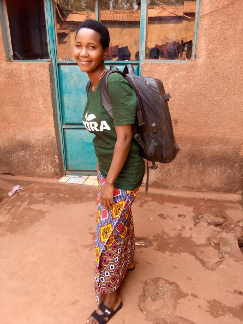 Young woman with short natural hair, smiling, wearing a green shirt with a partially visible white logo, colorful patterned skirt, black sandals, carrying a black backpack, standing outside in front of a textured orange wall with a weathered teal door and a glass window.