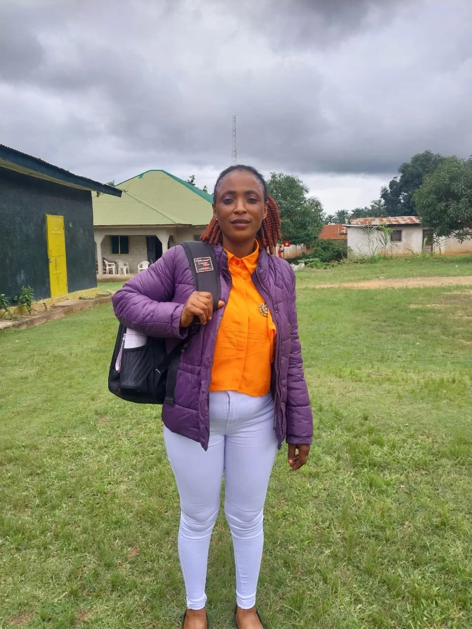 A woman standing outdoors on a grassy area, wearing an orange blouse, purple jacket, white pants, and carrying a black backpack, with dark clouds overhead and a few buildings and trees in the background.
