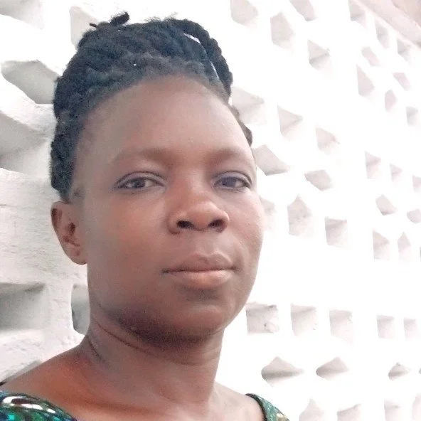 A woman with dark hair styled in twists, standing in front of a textured white wall with geometric cutouts.