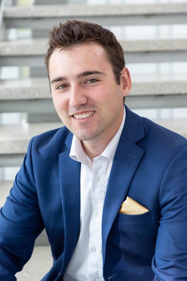 A person in a red T-shirt standing outdoors in a mountainous area with rocky terrain and pine trees.
