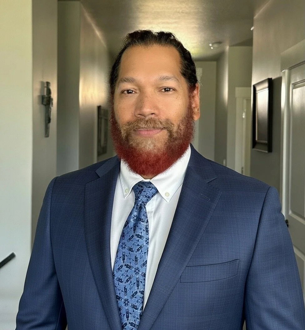 A man with a beard and mustache, wearing a striped shirt, looking at the camera with a slight smile.