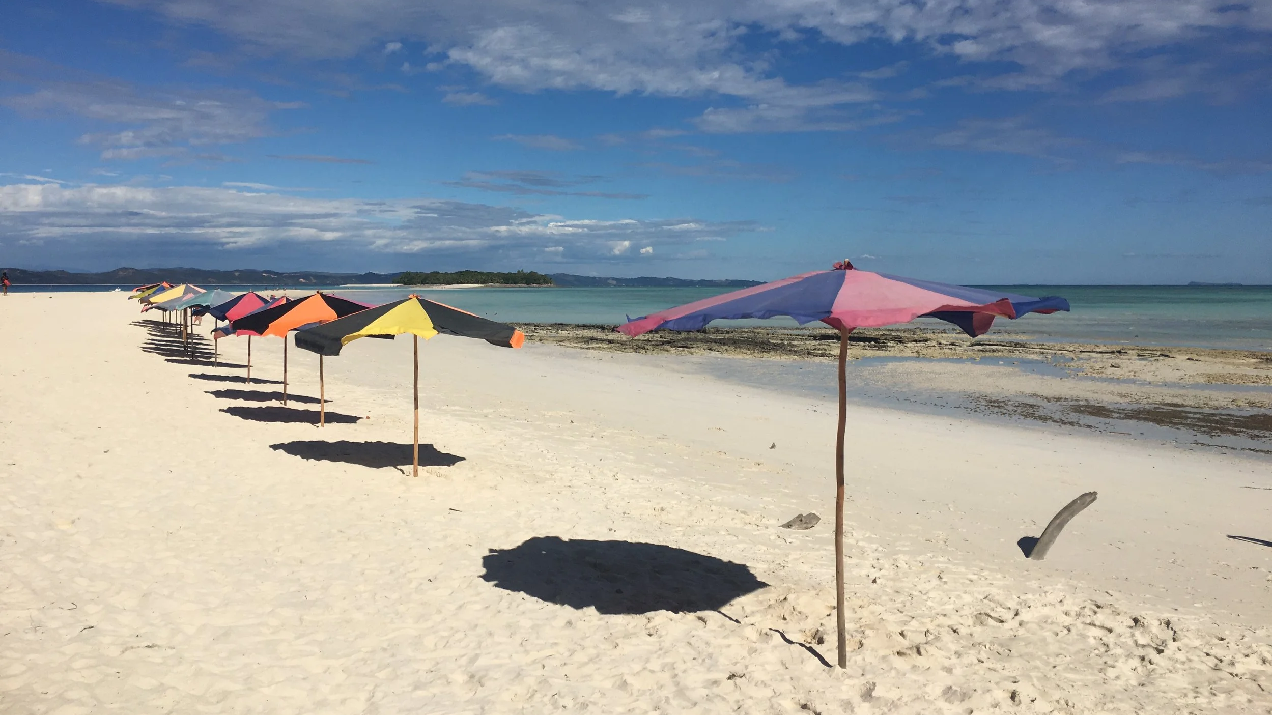 Colorful beach umbrellas lined up on a sandy beach near the water with a small island and cloudy sky in the background.