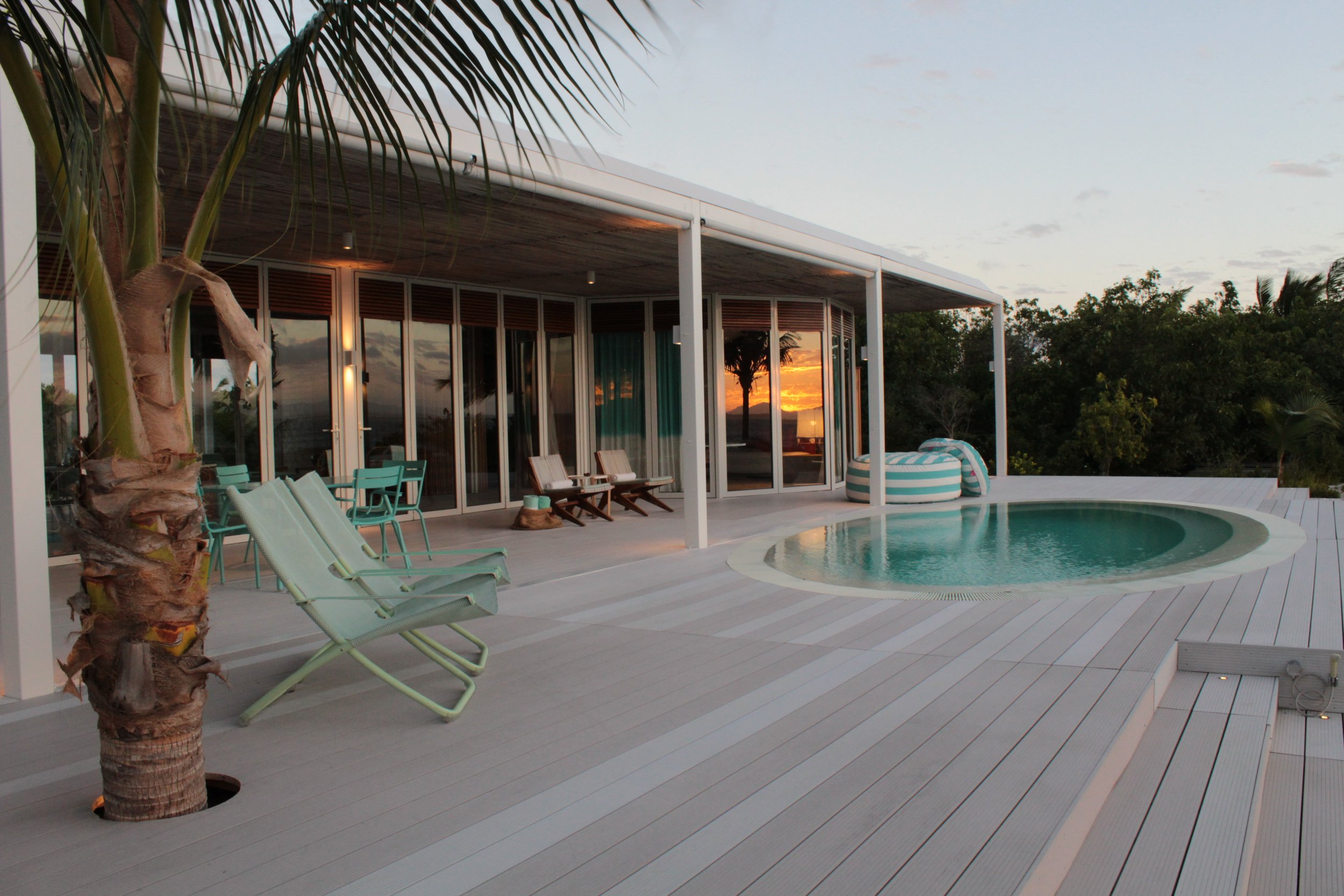A Miavana villa with a flat roof, large glass sliding doors, and an outdoor pool area at sunset. There are lounge chairs, a palm tree, and lush greenery surrounding the area.