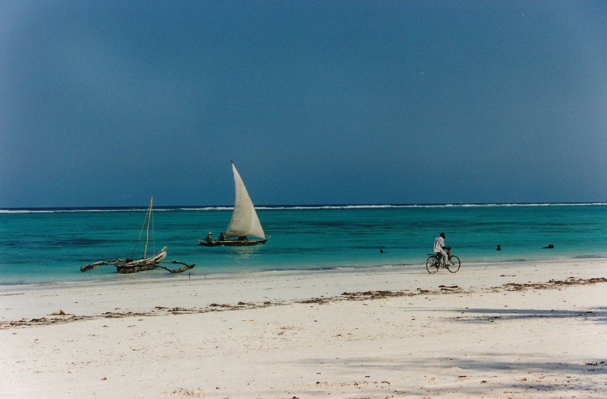 A man cycles along a beach with boats in the background