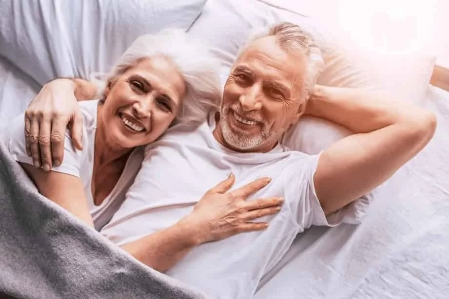 An elderly couple lying on a bed, smiling, with the woman hugging the man.