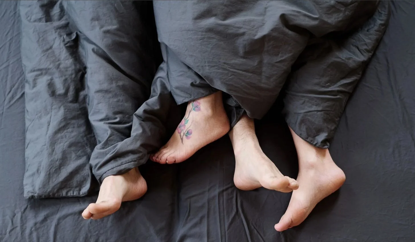 Two people lying on a bed intertwined, with their legs and feet visible. One person has a floral tattoo on their ankle.