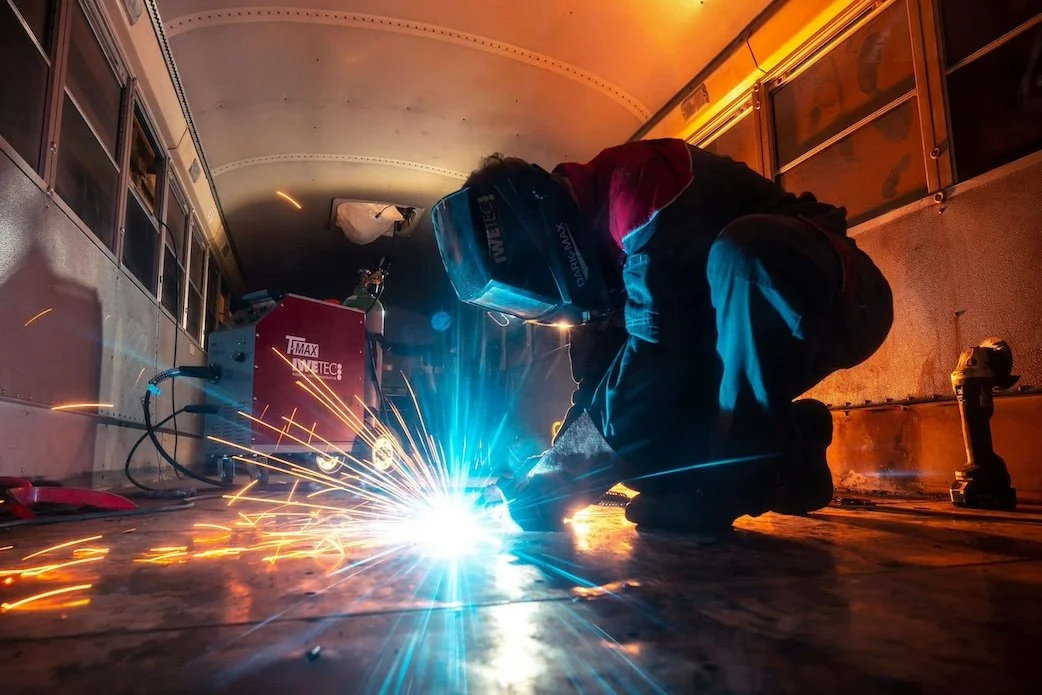 Person welding inside a metal enclosure, sparks flying, wearing protective helmet and gear.