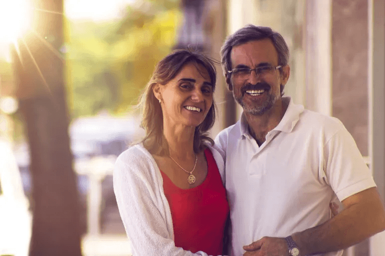 A middle-aged couple standing outdoors, smiling and embracing each other, with sunlight filtering through trees in the background.