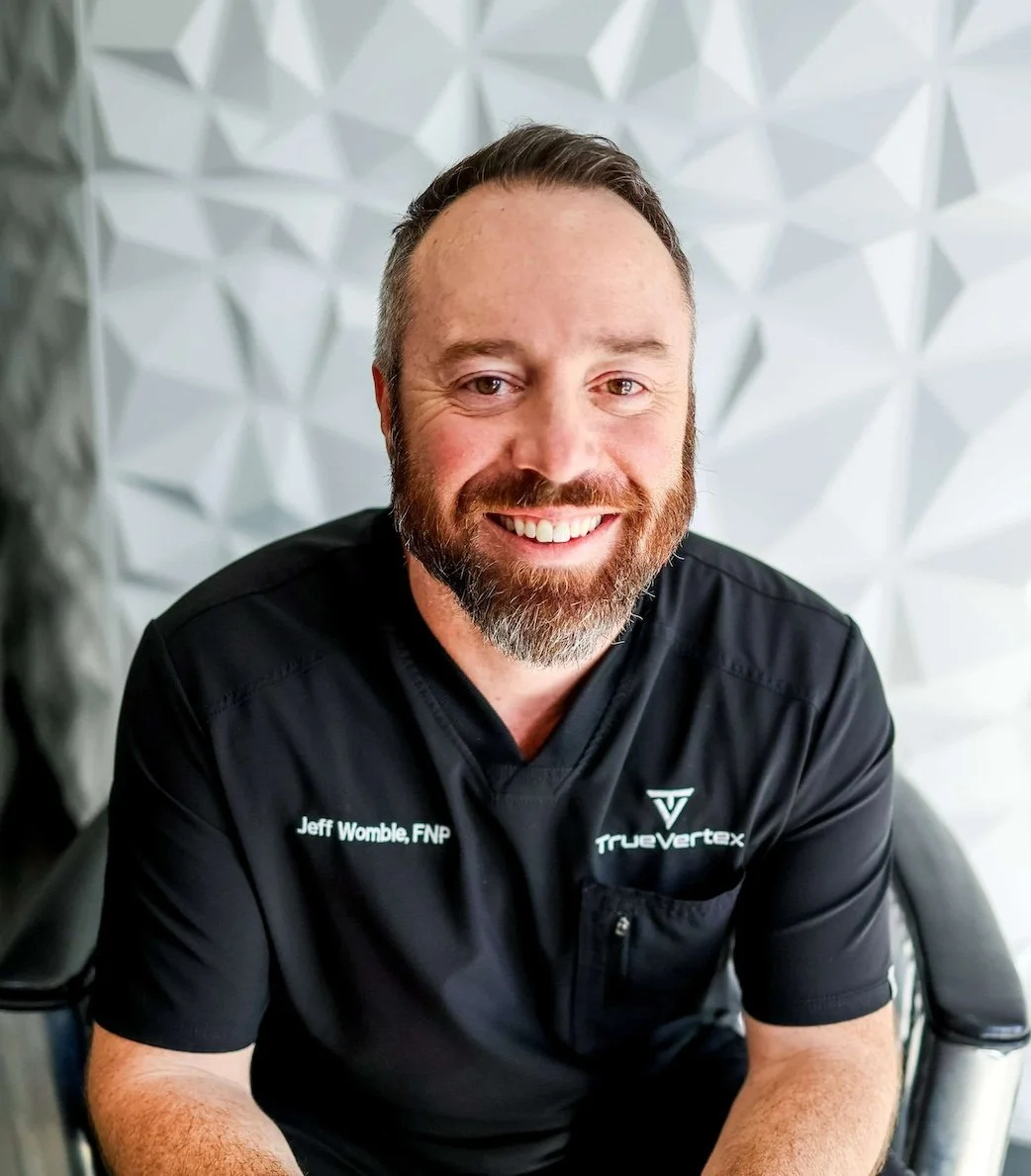 A smiling man with short dark hair and a beard, wearing a black medical uniform with 'Jeff Womble, FNP' and 'True Vertex' embroidery, sitting in a modern chair in front of a textured white wall.
