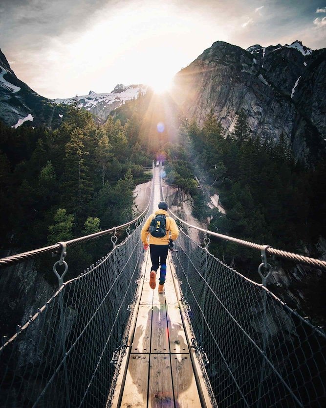 A person crossing a suspension bridge in a mountainous forest with snow-capped peaks and the sun shining through the clouds.