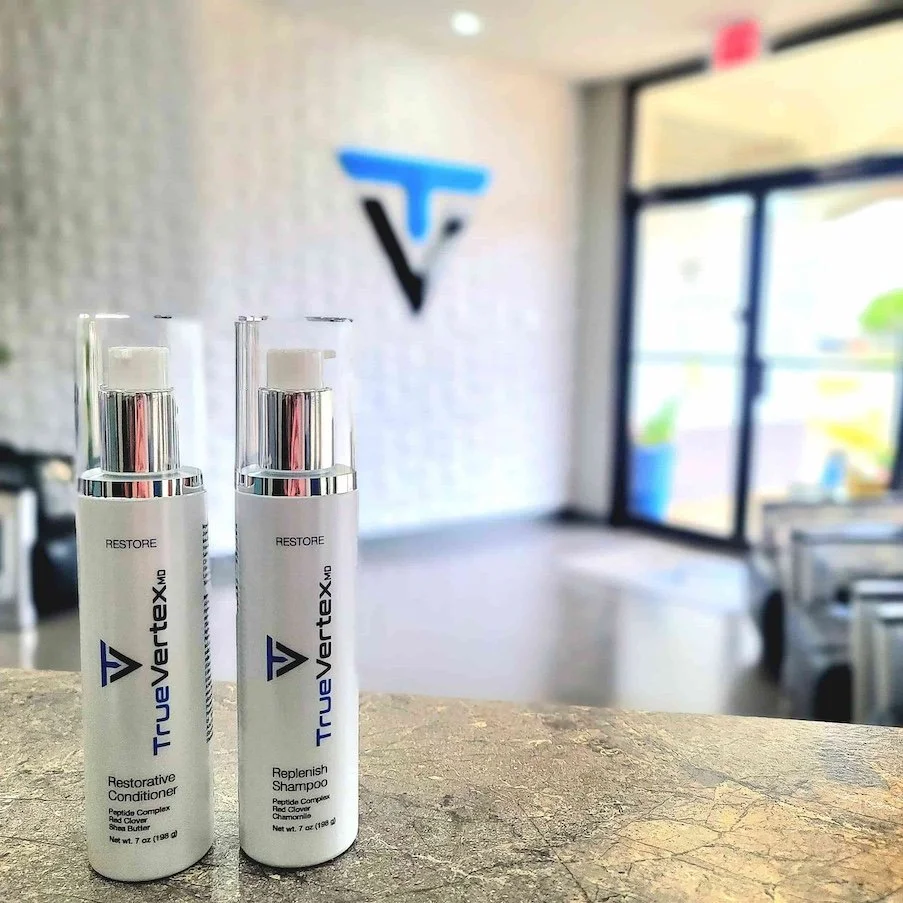 Two bottles of TrueVertaX MD hair care products, Restorative Conditioner and Replenish Shampoo, placed on a textured counter in a salon or spa setting with a blurred background showing a wall with a logo and large window.