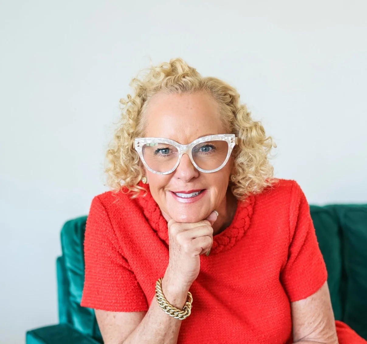 A woman with curly blonde hair, glasses, and a red top sitting on a teal chair.