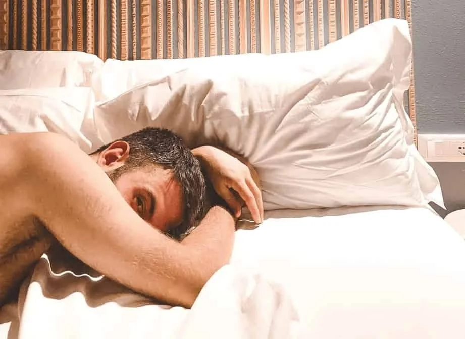 A shirtless man lying on a bed with white sheets, resting his head on a pillow, in a room with a wooden headboard and a gray wall.