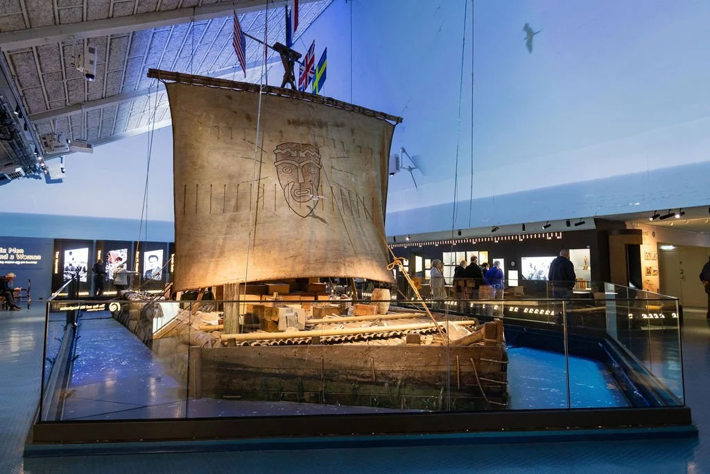 En utstilt historisk sjøfartøy med norske, britiske, svenske og amerikanske flagg i et museum.