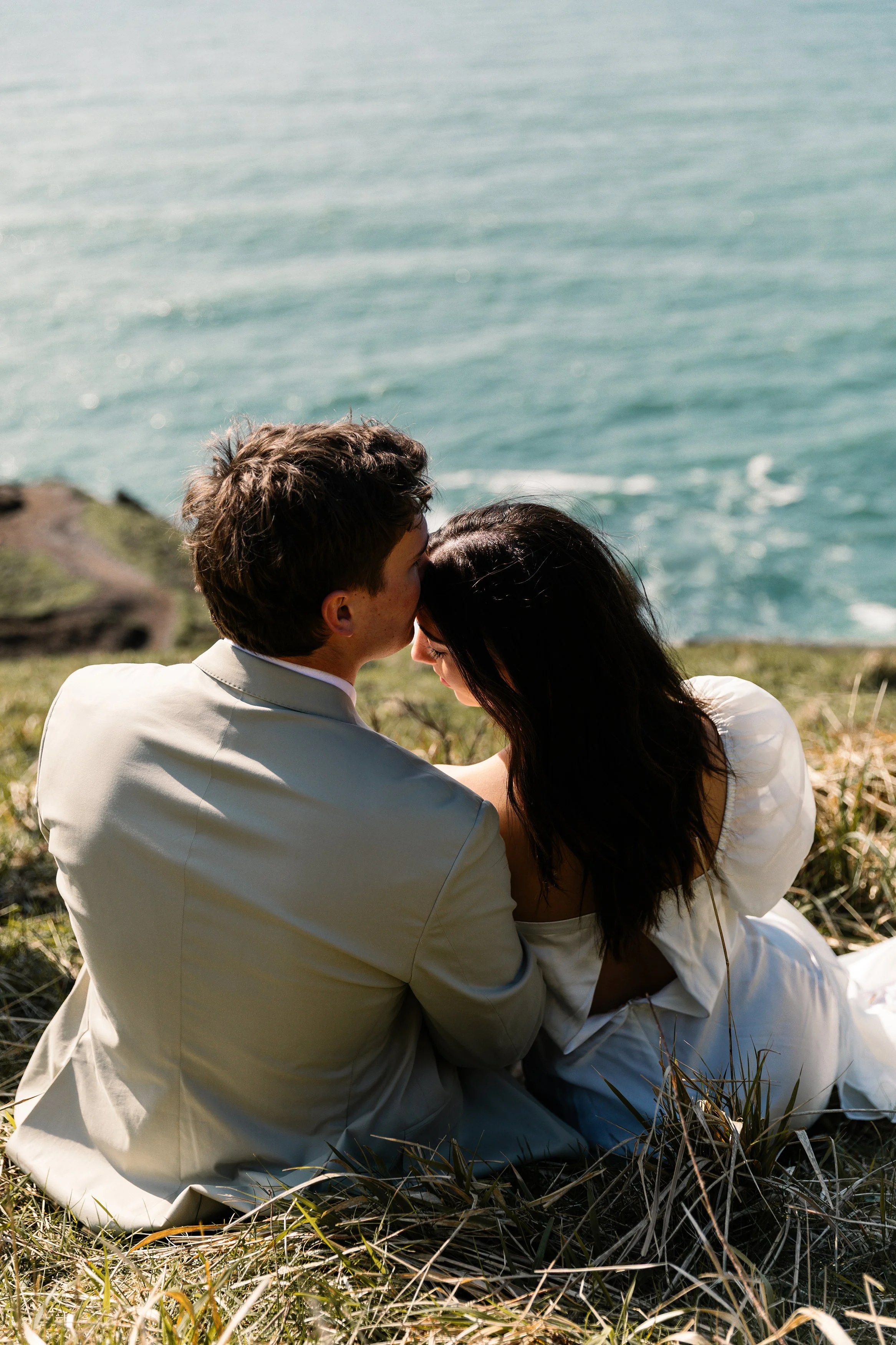 A couple sitting on a grassy coastal cliff, embracing and sharing a kiss with the ocean in the background. Elk Flats Oregon elopement