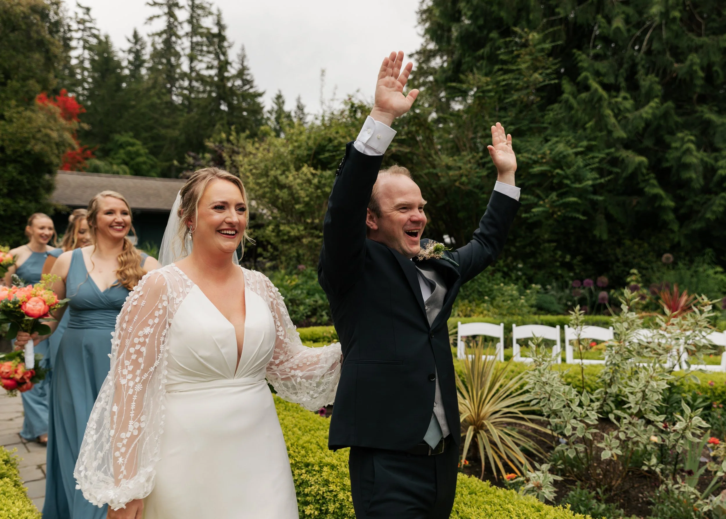A newlywed couple celebrating outdoors with bridesmaids in the background. The bride is wearing a white wedding dress with lace details and the groom is in a black suit with arms raised, smiling. The scene is set in a lush garden with trees and plant