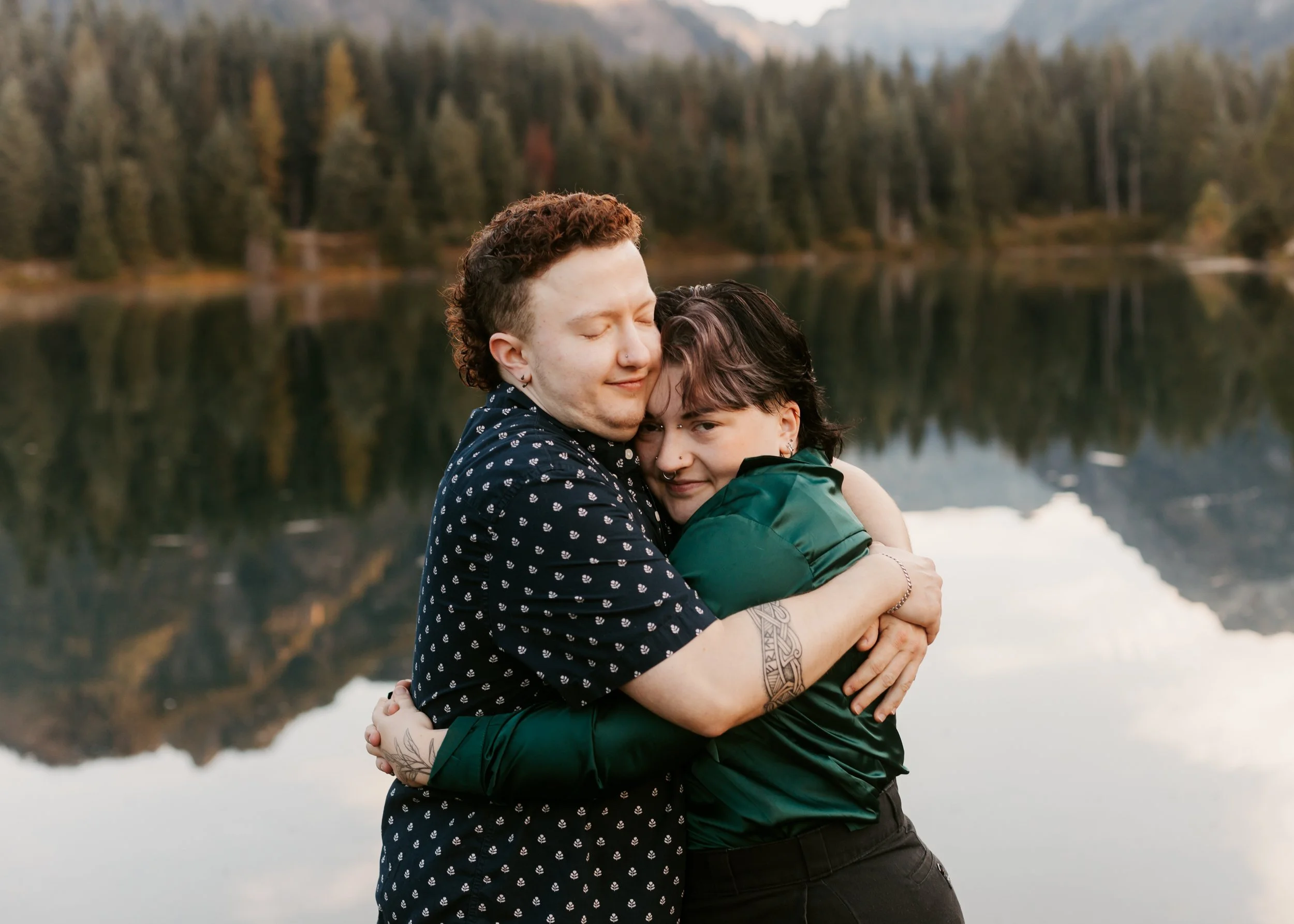 Two people hugging by a lake with trees and mountains in the background. Gold Creek Pond engagement