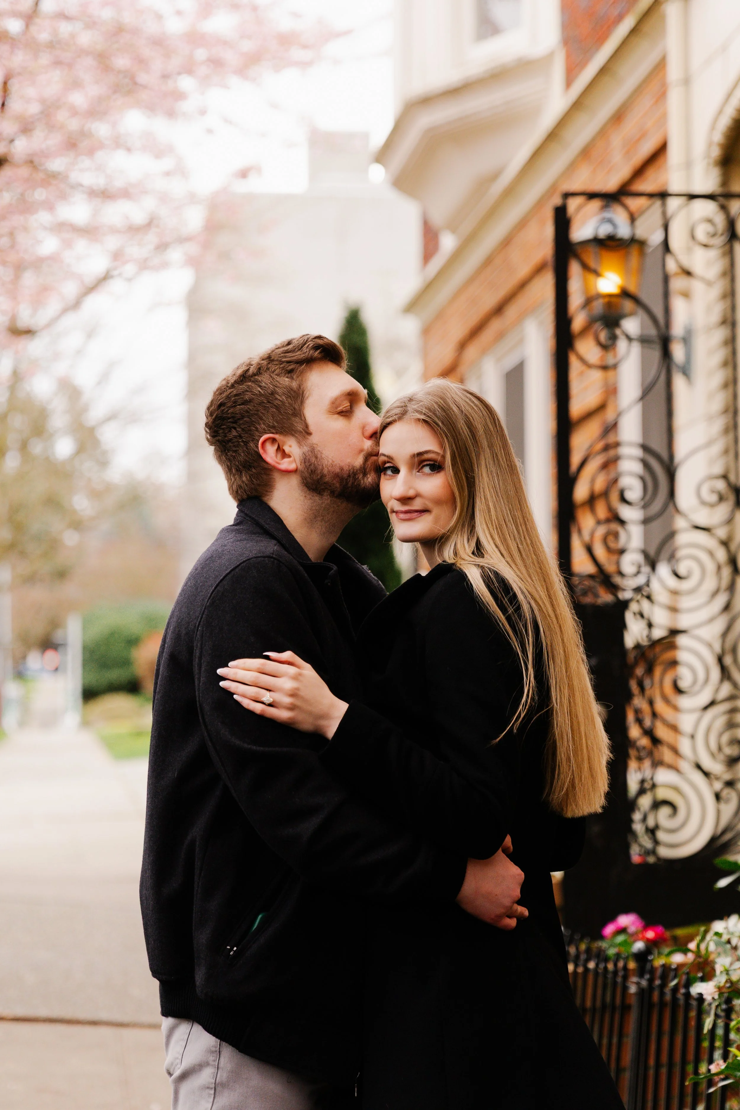 A couple embracing and sharing a kiss outdoors on a city sidewalk, with pink flowering trees and brick buildings in the background.