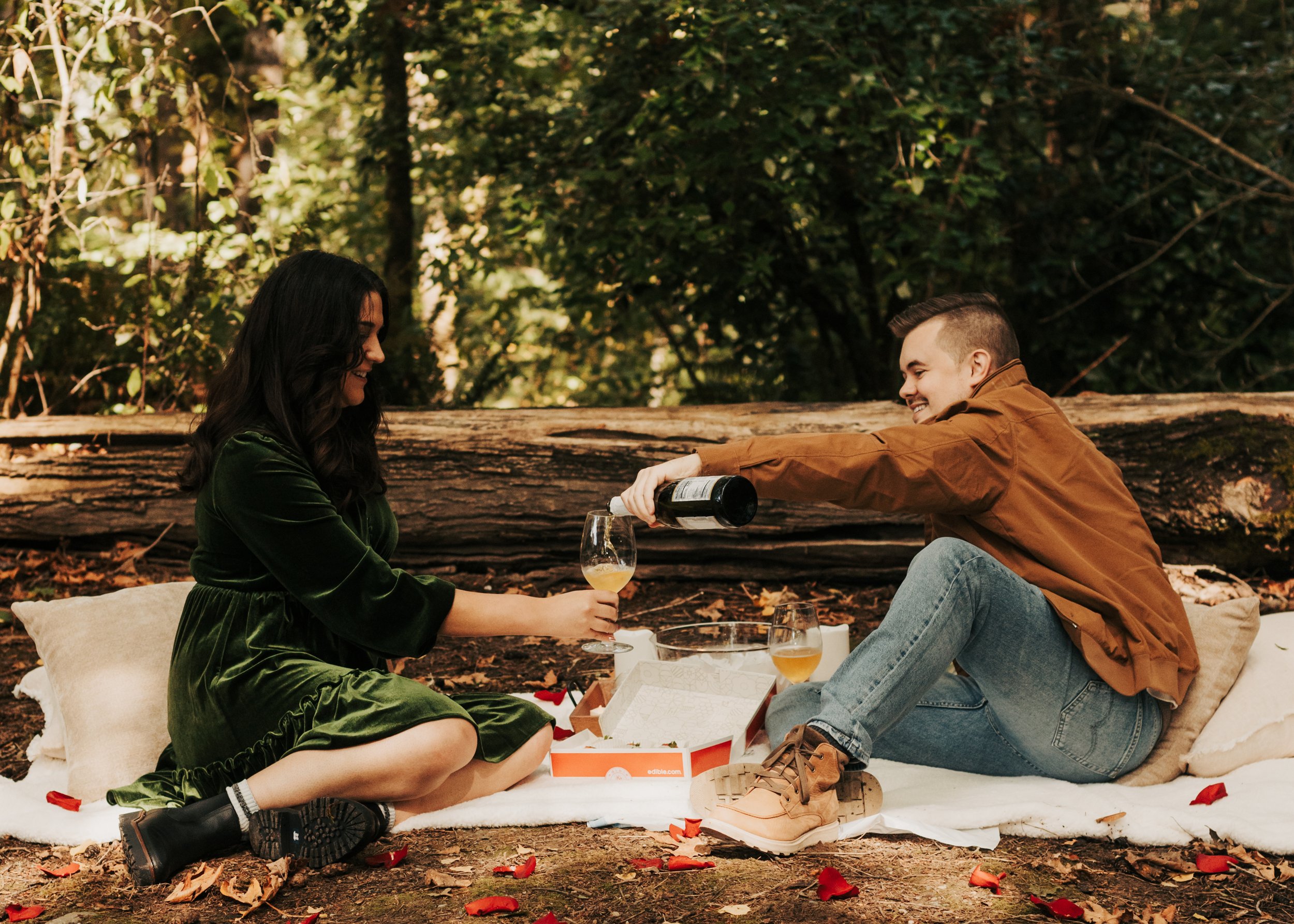 A man and woman sitting on a blanket in a forest, having a picnic. The man is pouring a drink into a glass held by the woman, and they are smiling at each other. St. Edwards State Park Washington engagement