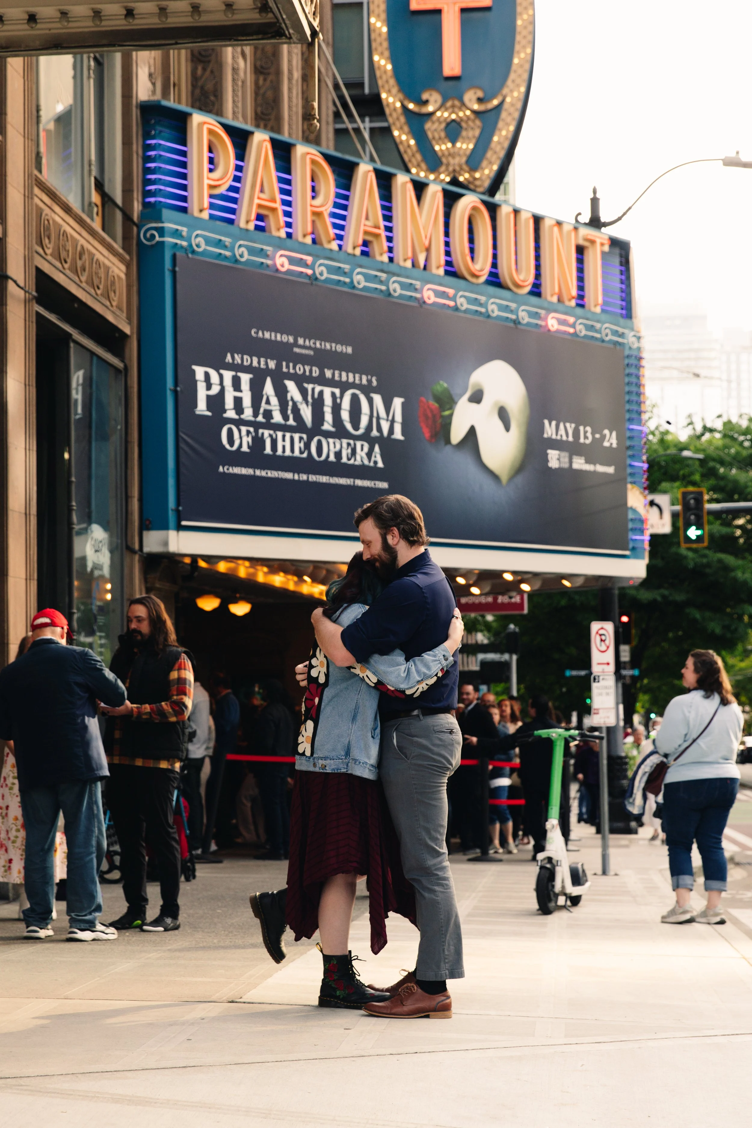 A couple hugging each other on a city sidewalk in front of a movie theater marquee for "Phantom of the Opera" in an urban setting with other people walking and standing around.