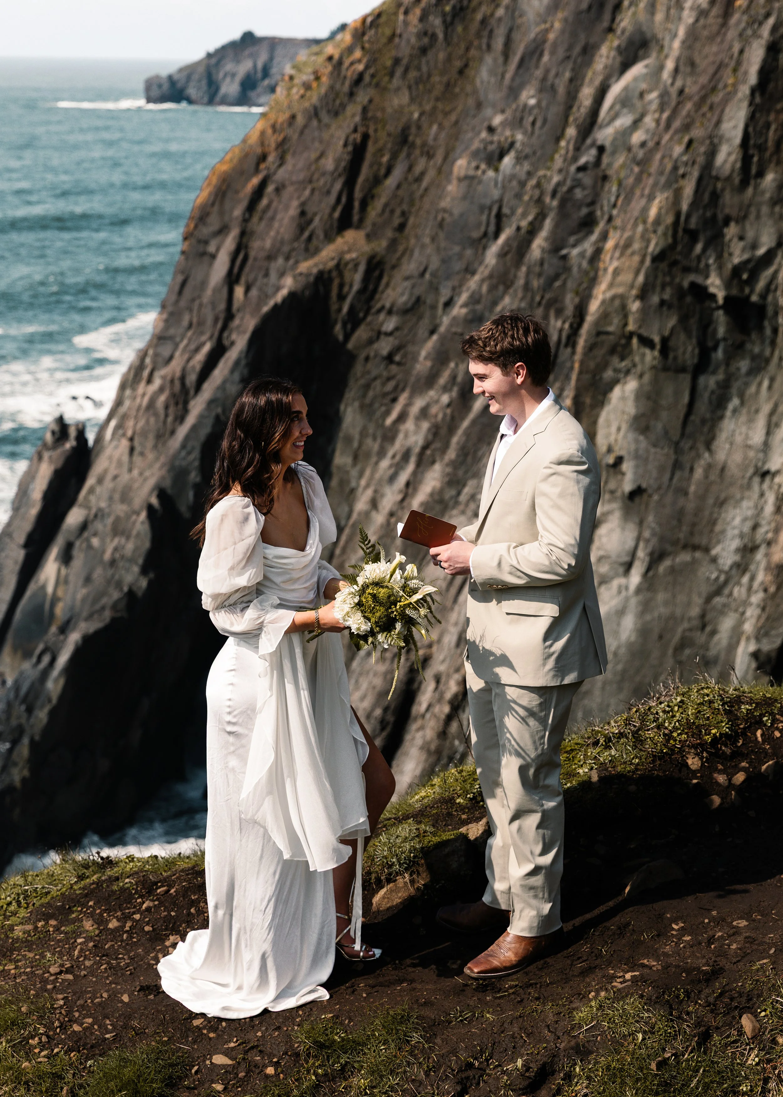 A couple getting eloped outdoors on a rocky cliffside near the ocean, with the bride holding a bouquet and the groom reading vows. Elk Flats Oregon