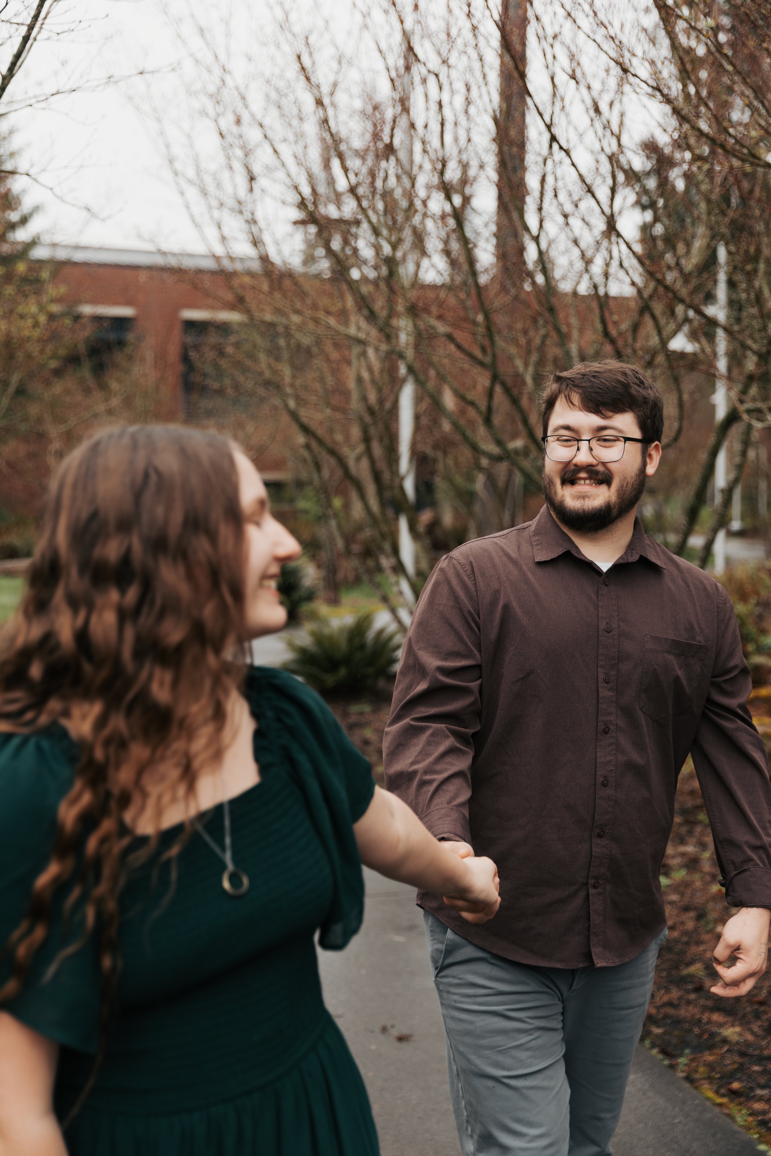Two people holding hands and walking outdoors on a cloudy day, with trees and a building in the background.
