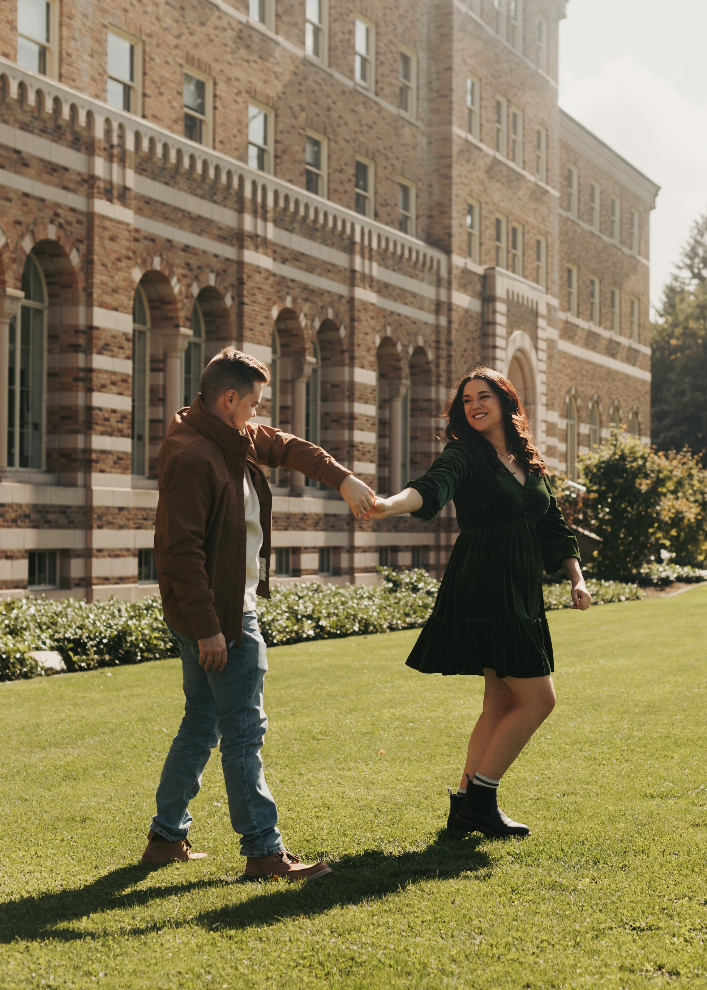 A young man and woman holding hands and dancing on a lush green lawn in front of a large brick building with multiple windows. The woman is smiling and wearing a black dress and boots, while the man is dressed casually in a brown jacket and jeans. Th