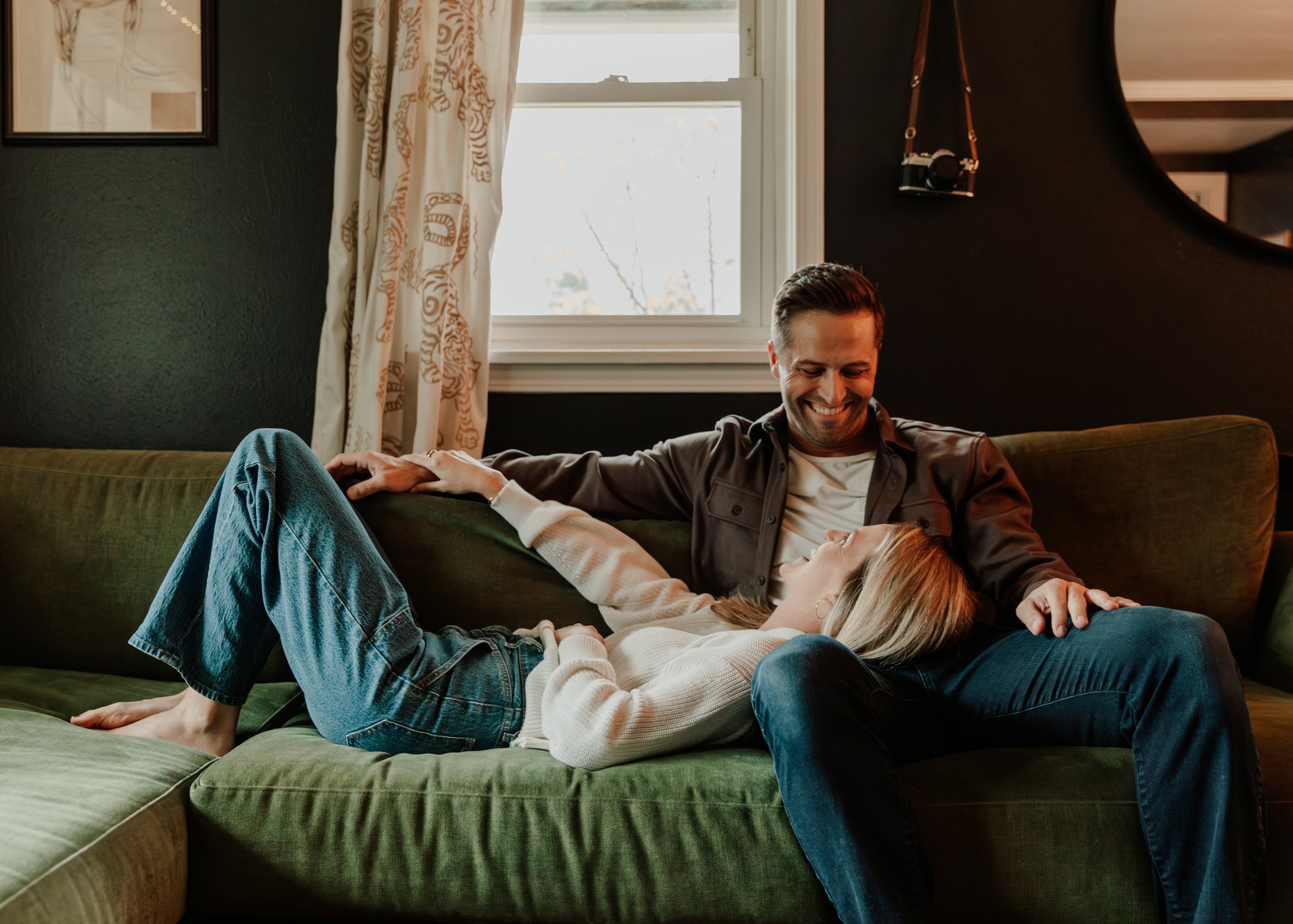 A man and woman sitting on a green couch in a cozy living room, smiling and enjoying each other's company, with a large window and curtains behind them. Indoor couples photography
