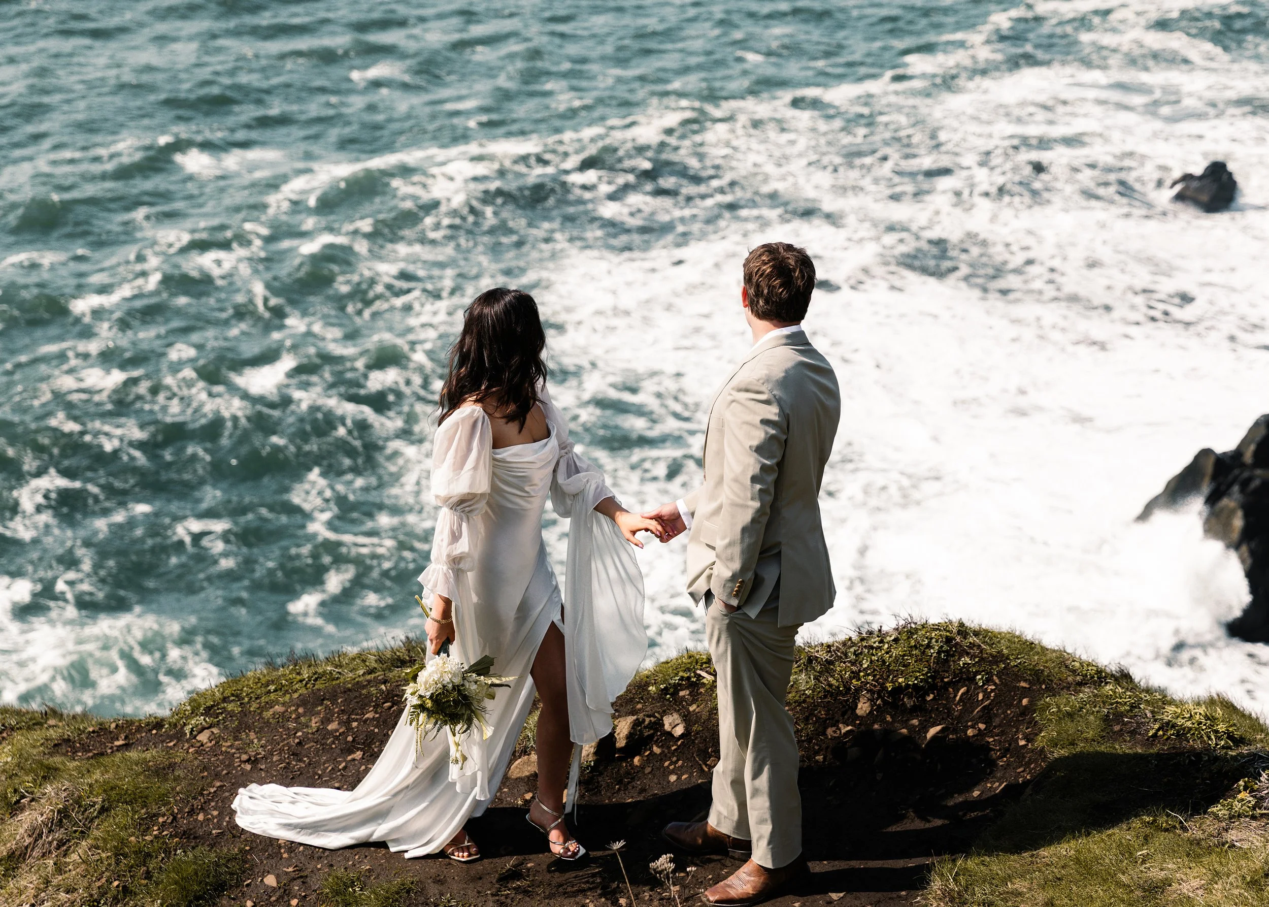 A bride and groom standing on a grassy cliff by the ocean, holding hands during their wedding ceremony with waves crashing in the background. Elk Flats Oregon