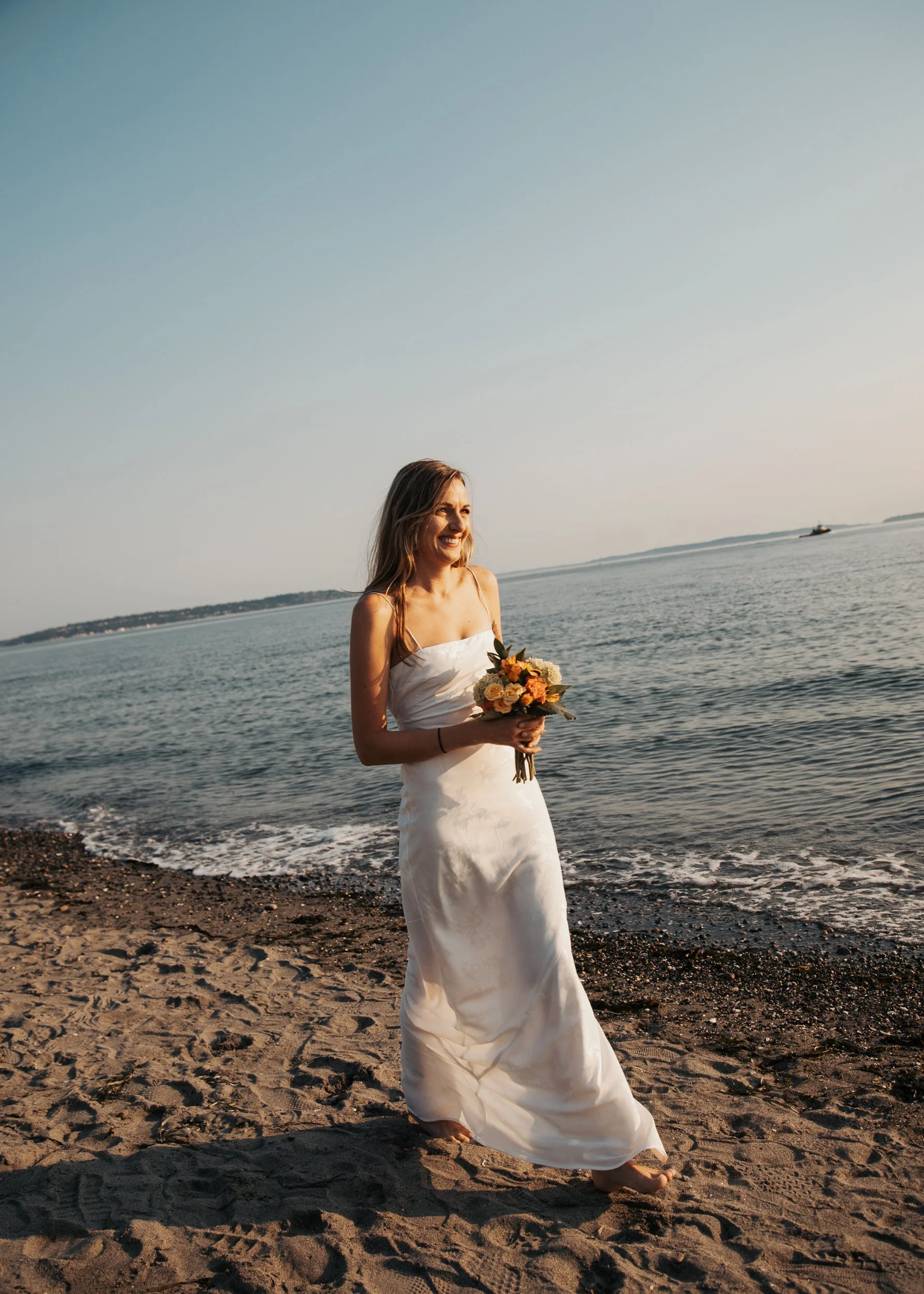 A woman in a white dress holding a bouquet of flowers walking barefoot on a beach during sunset. Discovery Park Beach Lighthouse elopement
