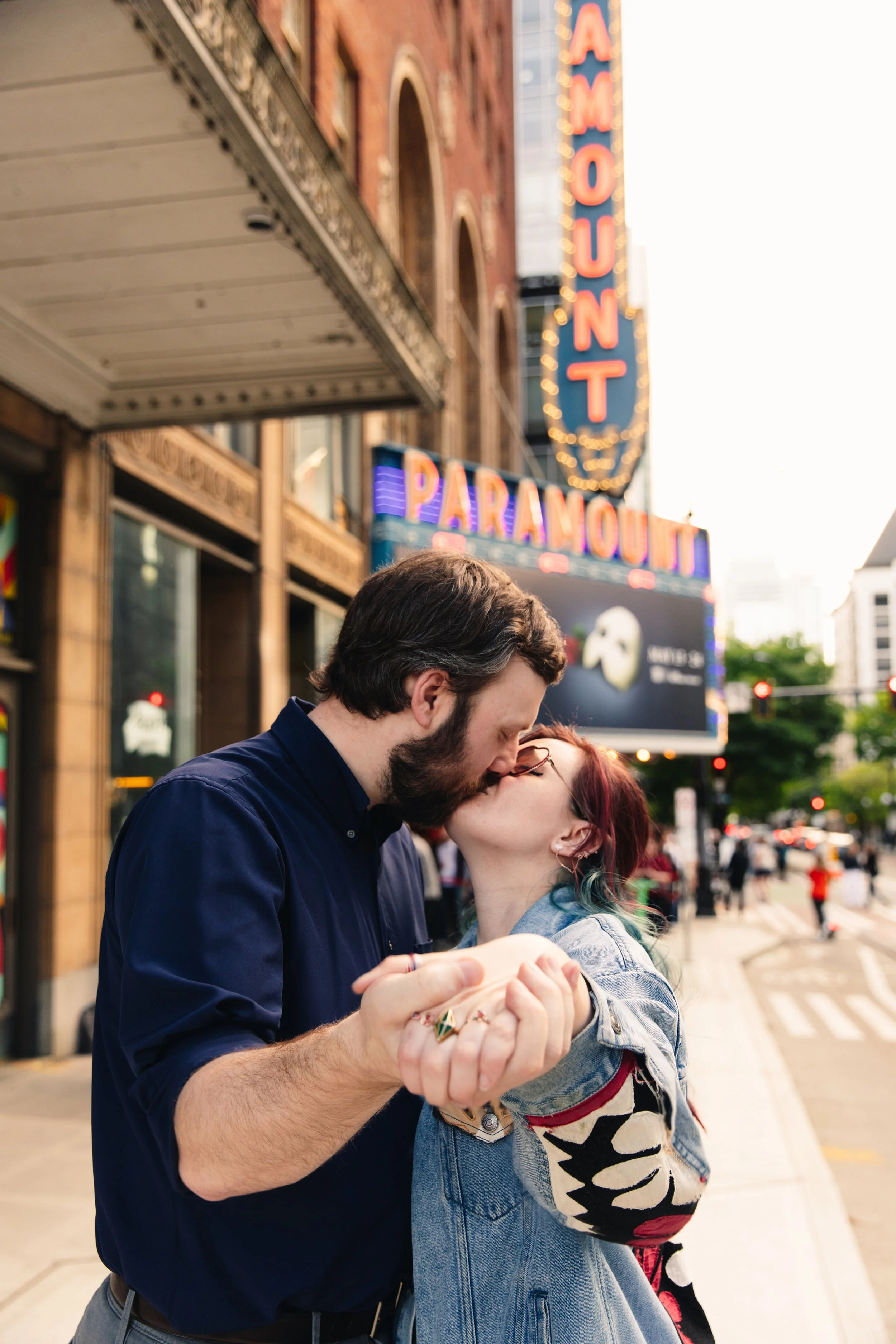 A man and a woman sharing a kiss on a busy street with neon signs for the Paramount Theatre in the background. The man is wearing a navy blue shirt and has a beard, and the woman has colorful hair and glasses.