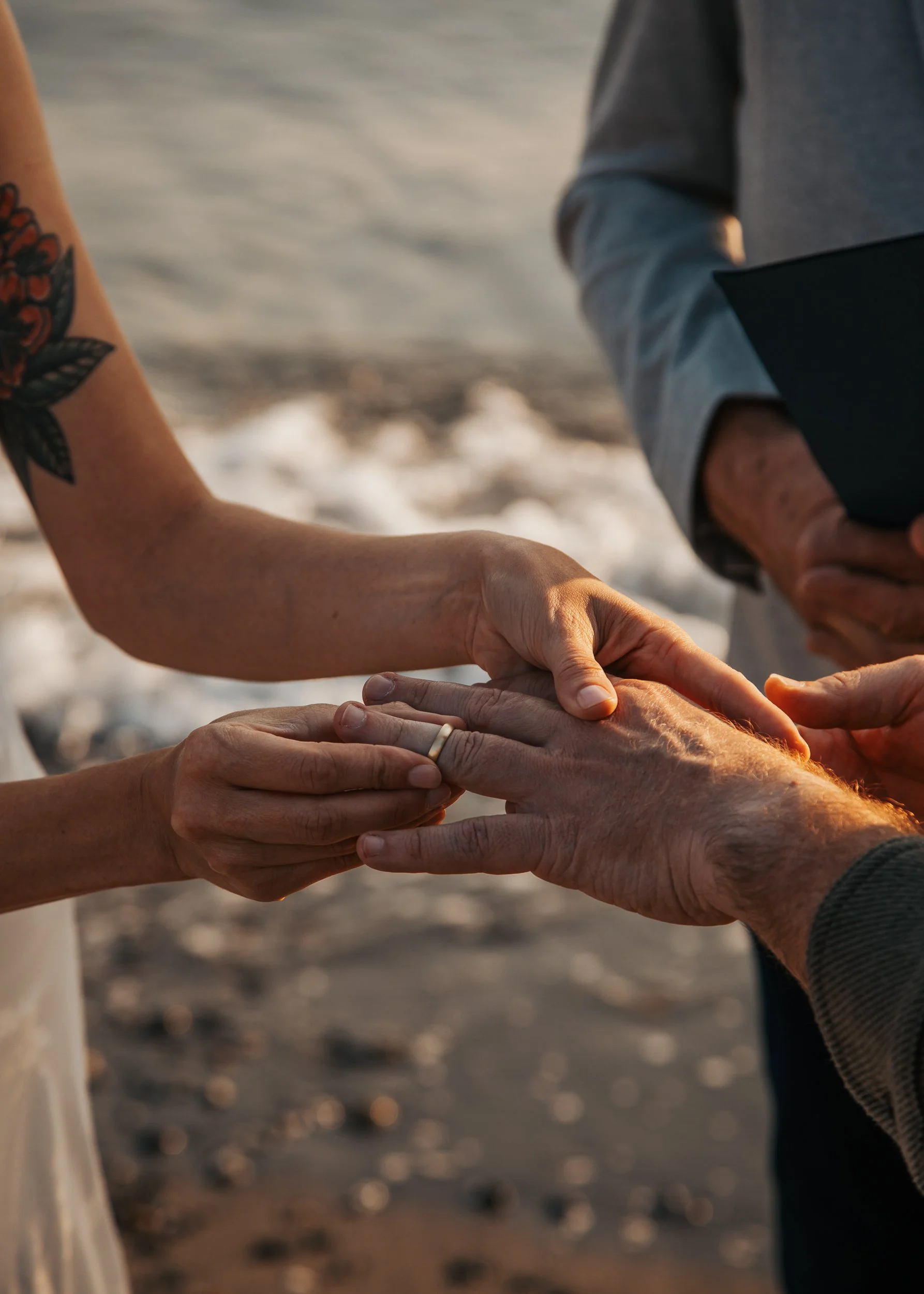 A couple exchanging rings during a wedding on a beach at sunset, with one person holding another's hand. Discovery Park Beach Lighthouse elopement