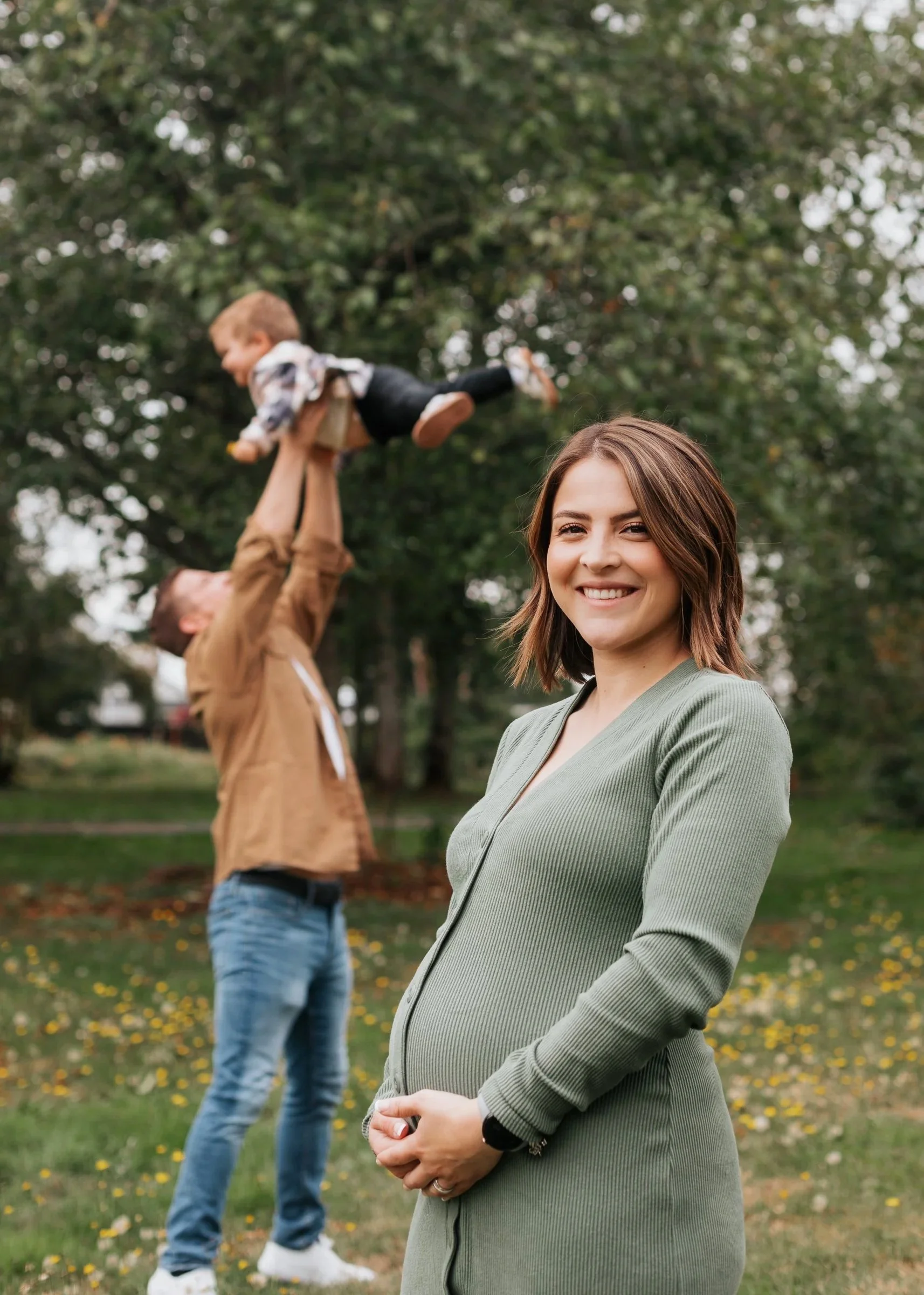 A pregnant woman smiling outdoors with a man in the background lifting a young boy in the air. The scene is in Nelson Nature Park in Washington state.