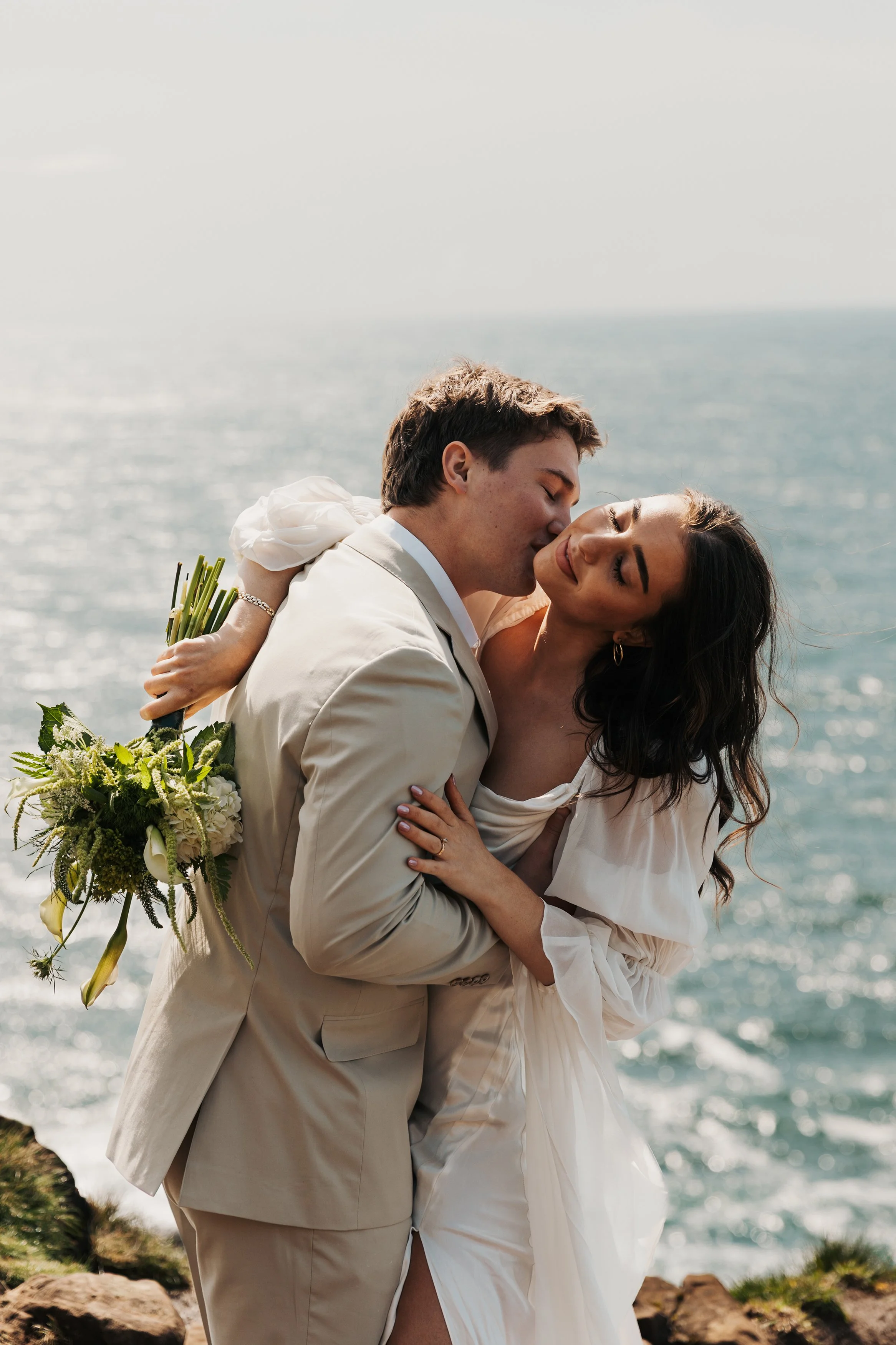 A couple in wedding attire embracing on a rocky coastline with the ocean in the background, the man kissing the woman's cheek. Elk Flats Oregon elopement