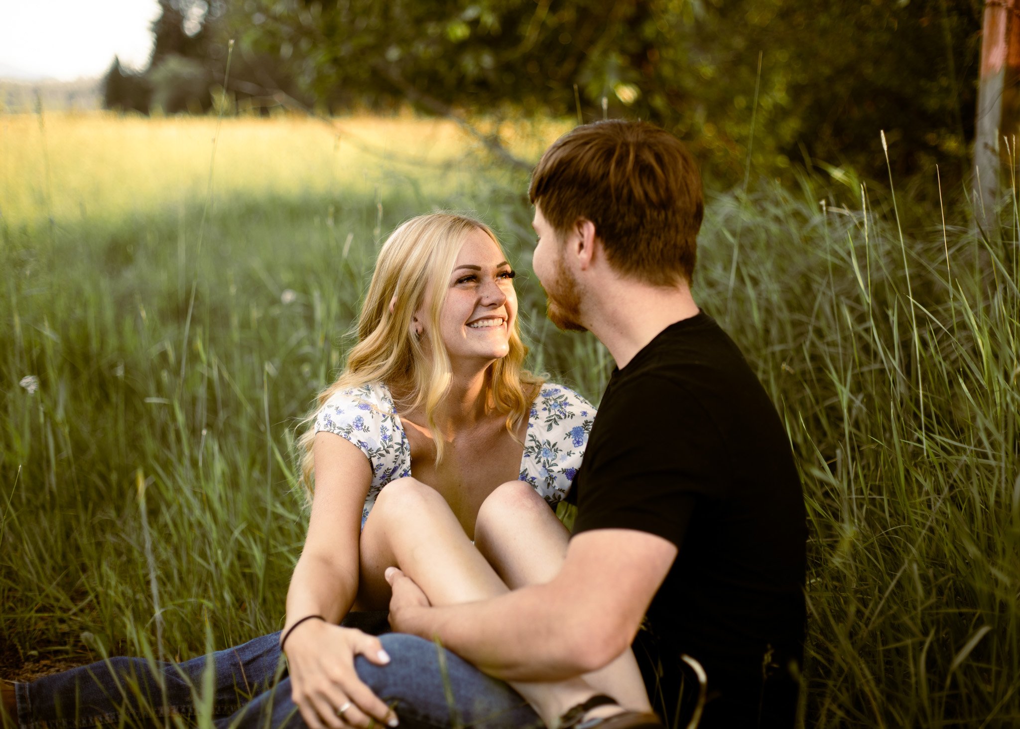 A couple sits in a grassy field, smiling and looking into each other's eyes, surrounded by tall grass and trees in the background.