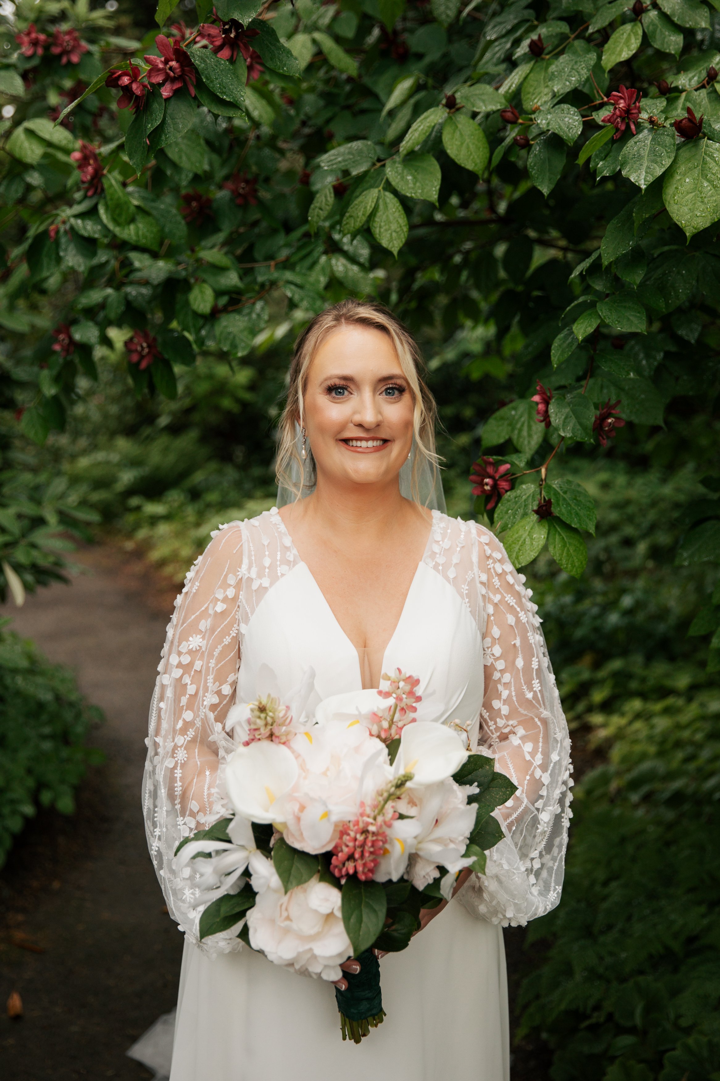 A bride in a white wedding dress holding a bouquet of white and pink flowers outdoors surrounded by lush green foliage. Heronswood Garden wedding Kingston Washington