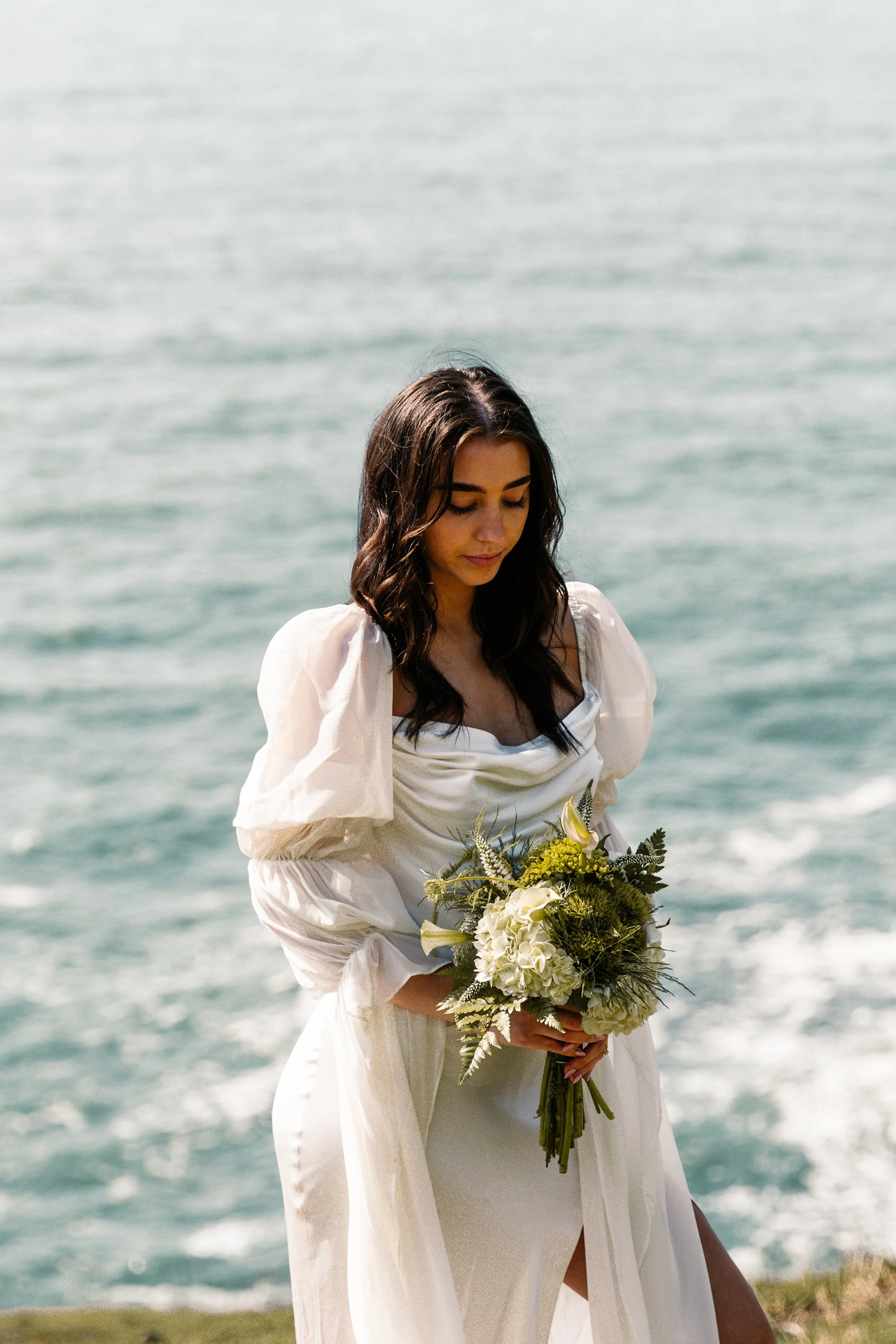 A woman in a white dress holding a bouquet of white and yellow flowers standing near the water with an ocean in the background. Elk Flats Oregon elopement
