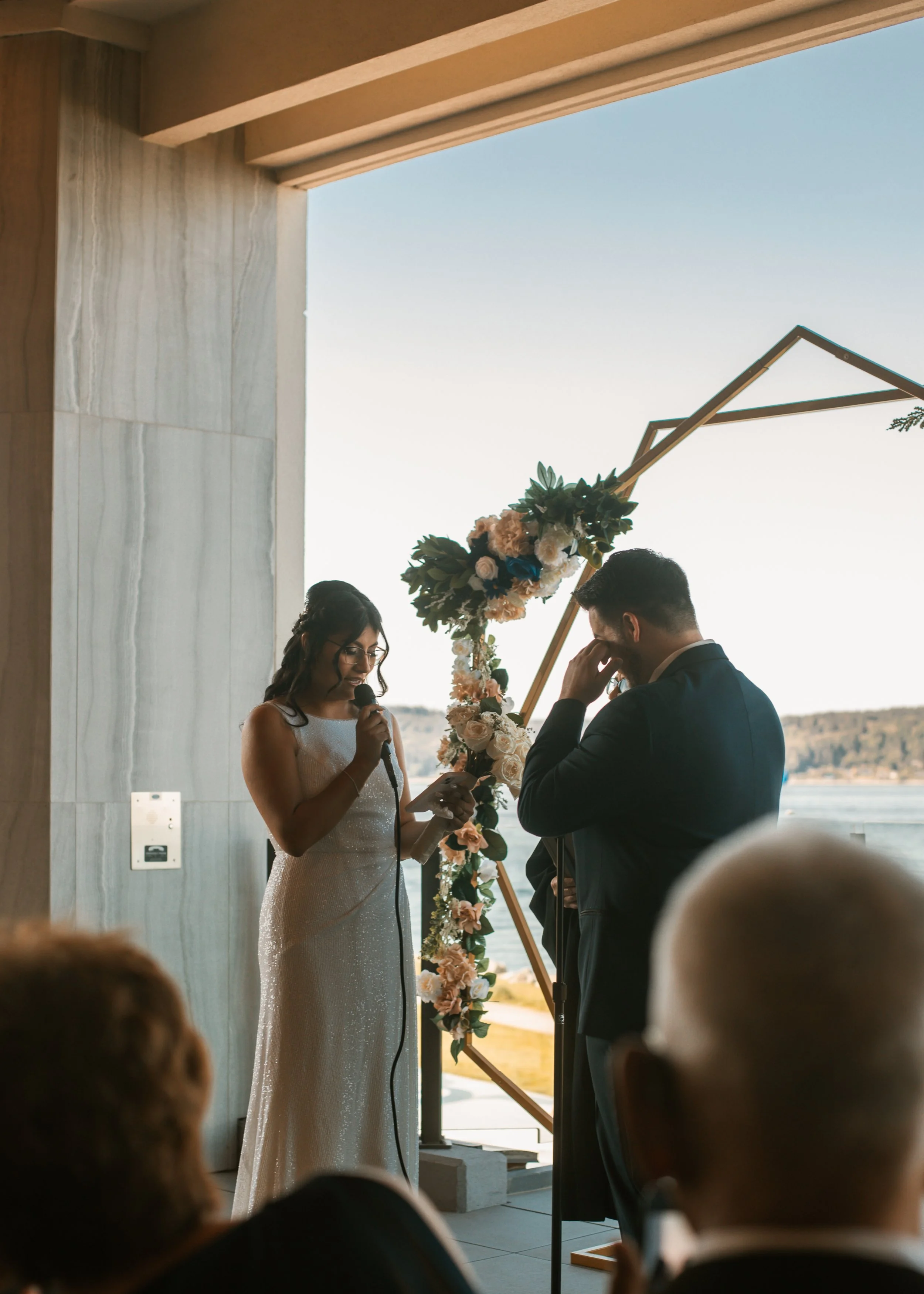 A woman in a white wedding dress reads vows into a microphone while a man in a dark suit wipes tears during their wedding ceremony, with an outdoor scenic view in the background.
