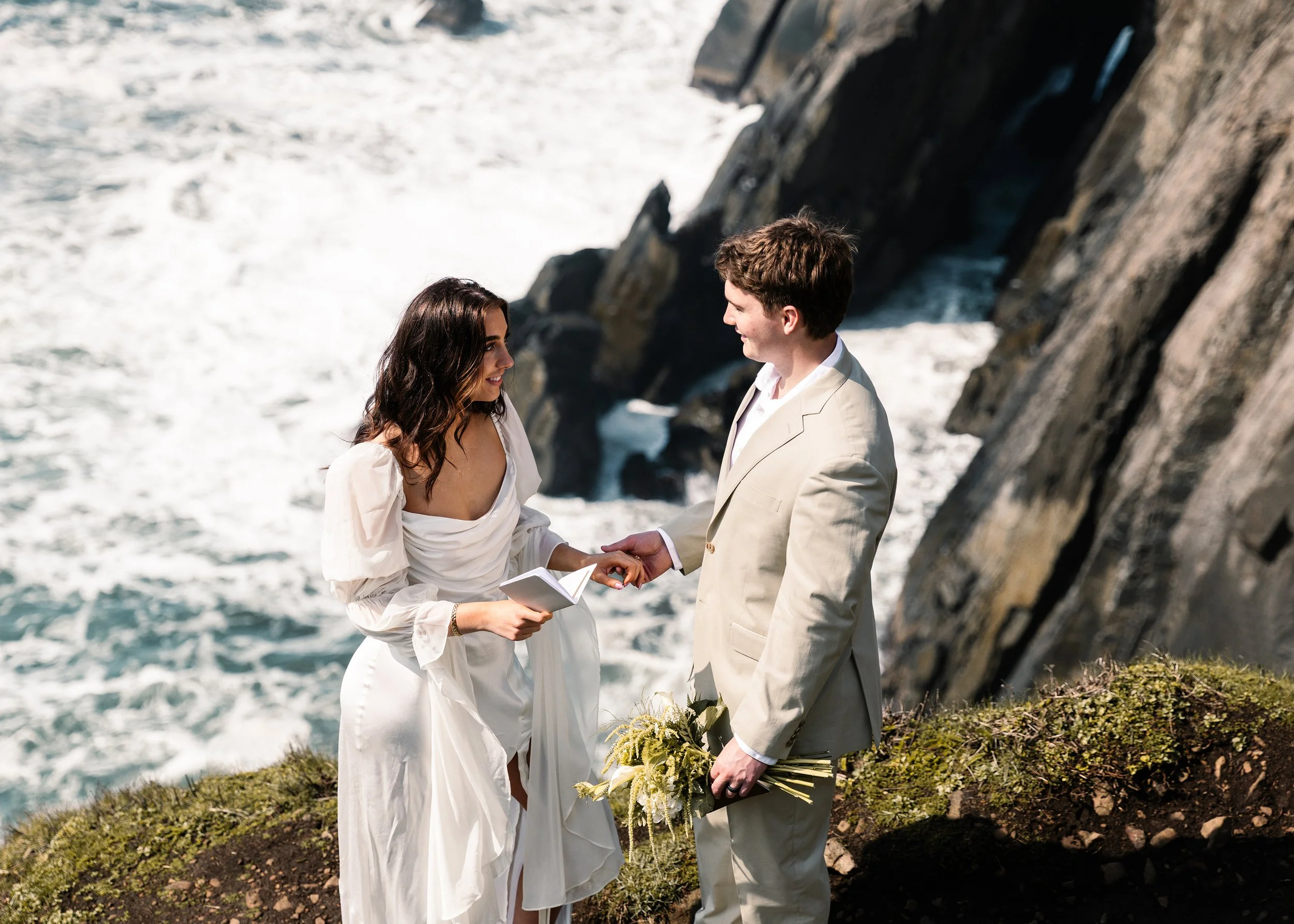 A couple exchanging vows during their wedding ceremony on a rocky coastal cliff, with the ocean in the background. Elk Flats Oregon