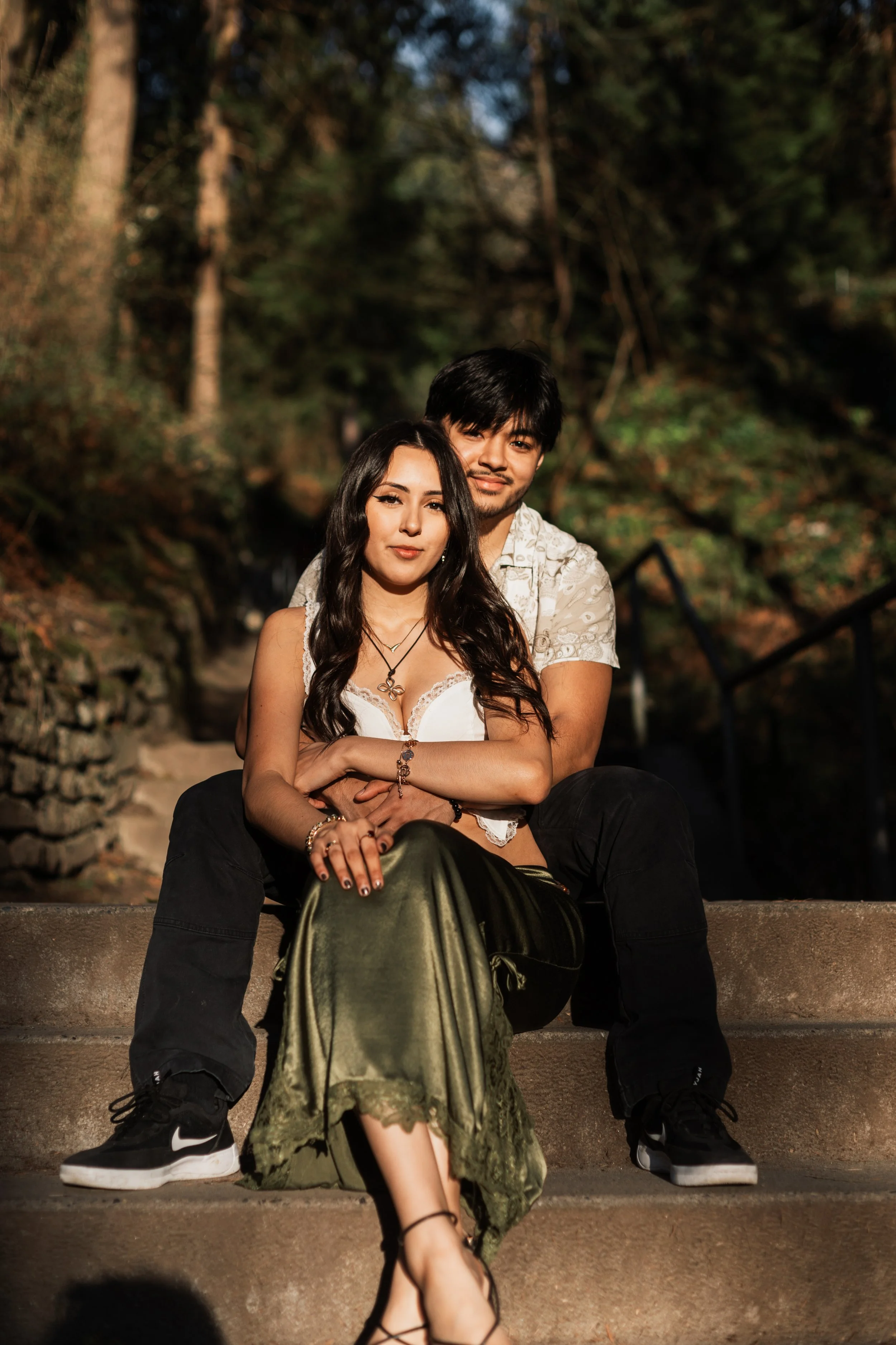 A young couple sitting on outdoor steps with greenery in the background, embracing and looking at the camera. Golden Gardens park engagement photos