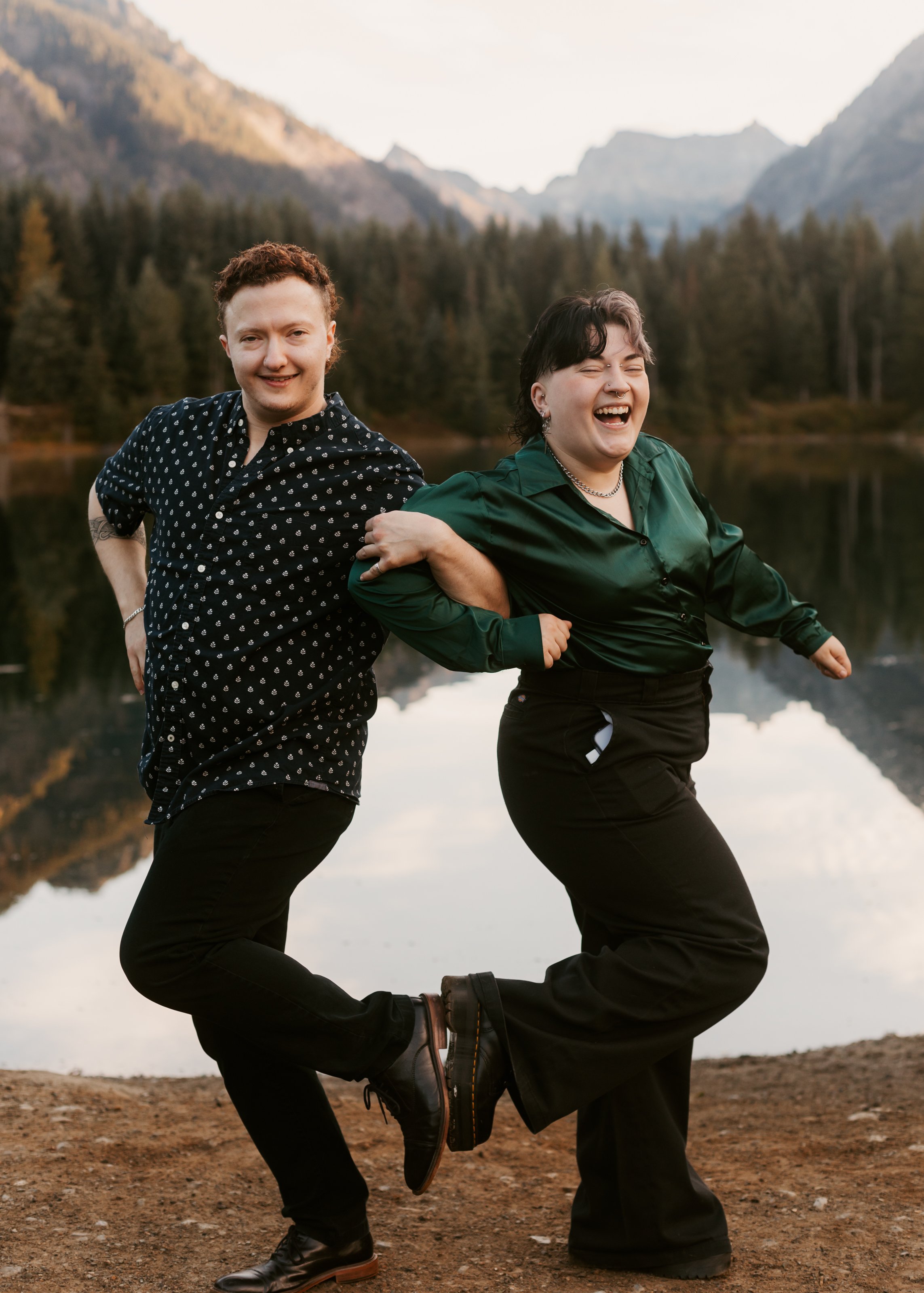 Two people dancing by a lake in a mountain landscape, one with curly hair and the other with short dark hair, both smiling and holding each other. Gold Creek Pond engagement