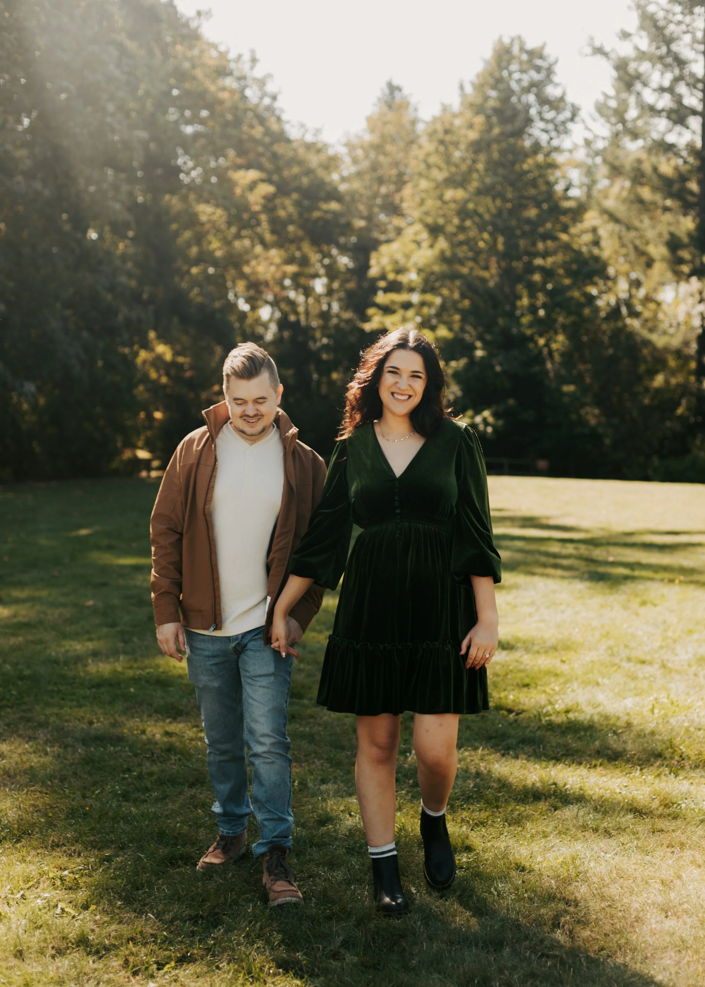 A smiling woman and a man walking hand in hand outdoors in a park with fall foliage, sunlight filtering through trees, and grassy ground.