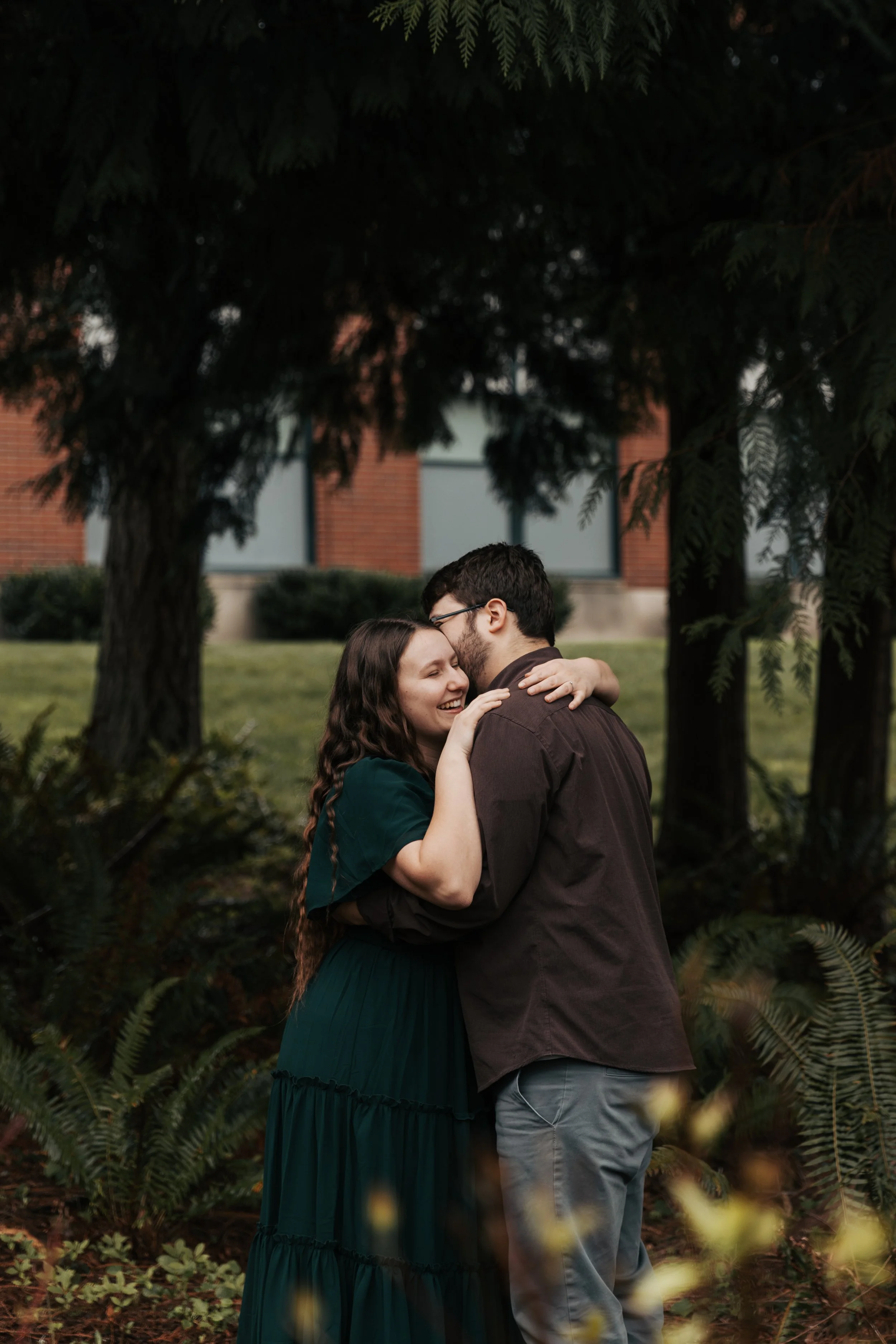 A couple embracing outdoors in front of a large tree and greenery, with a building with brick and windows in the background.