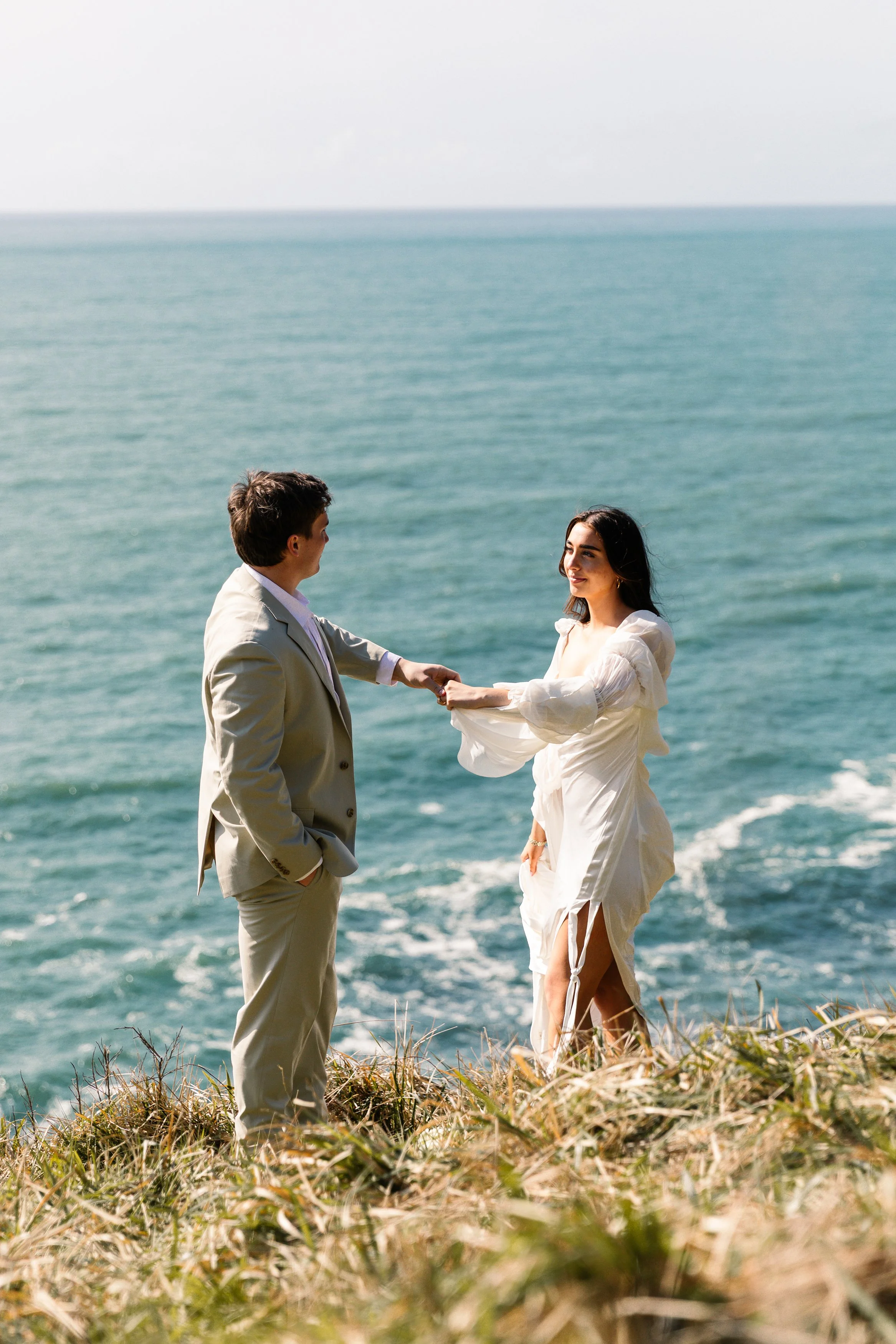 A couple dressed in wedding attire standing on a grassy cliff overlooking the ocean, holding hands. Elk Flats Oregon elopement