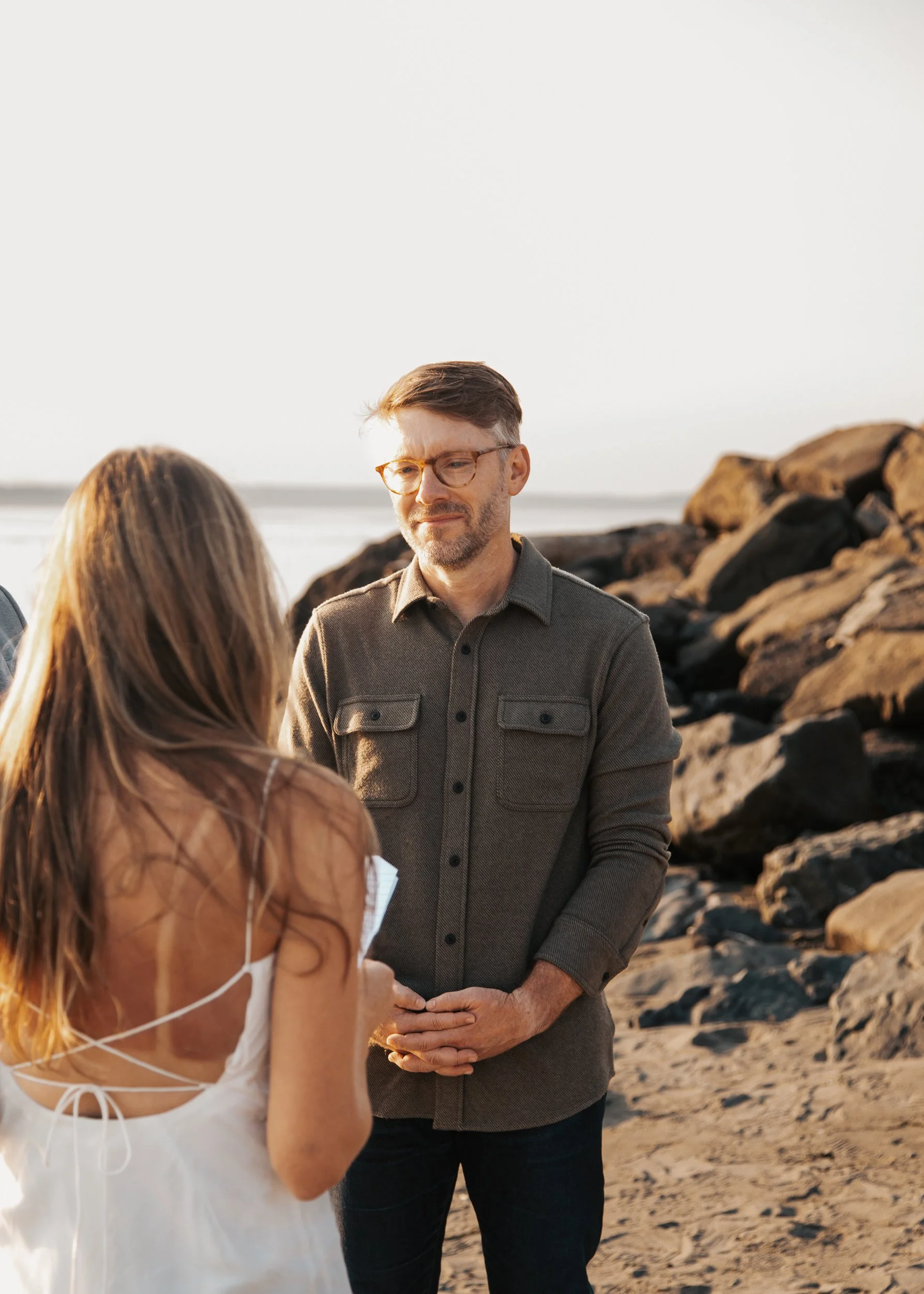 A man and woman standing on a beach, facing each other. The man is wearing glasses and a gray shirt, and there are rocks in the background. Discovery Park Beach Lighthouse elopement