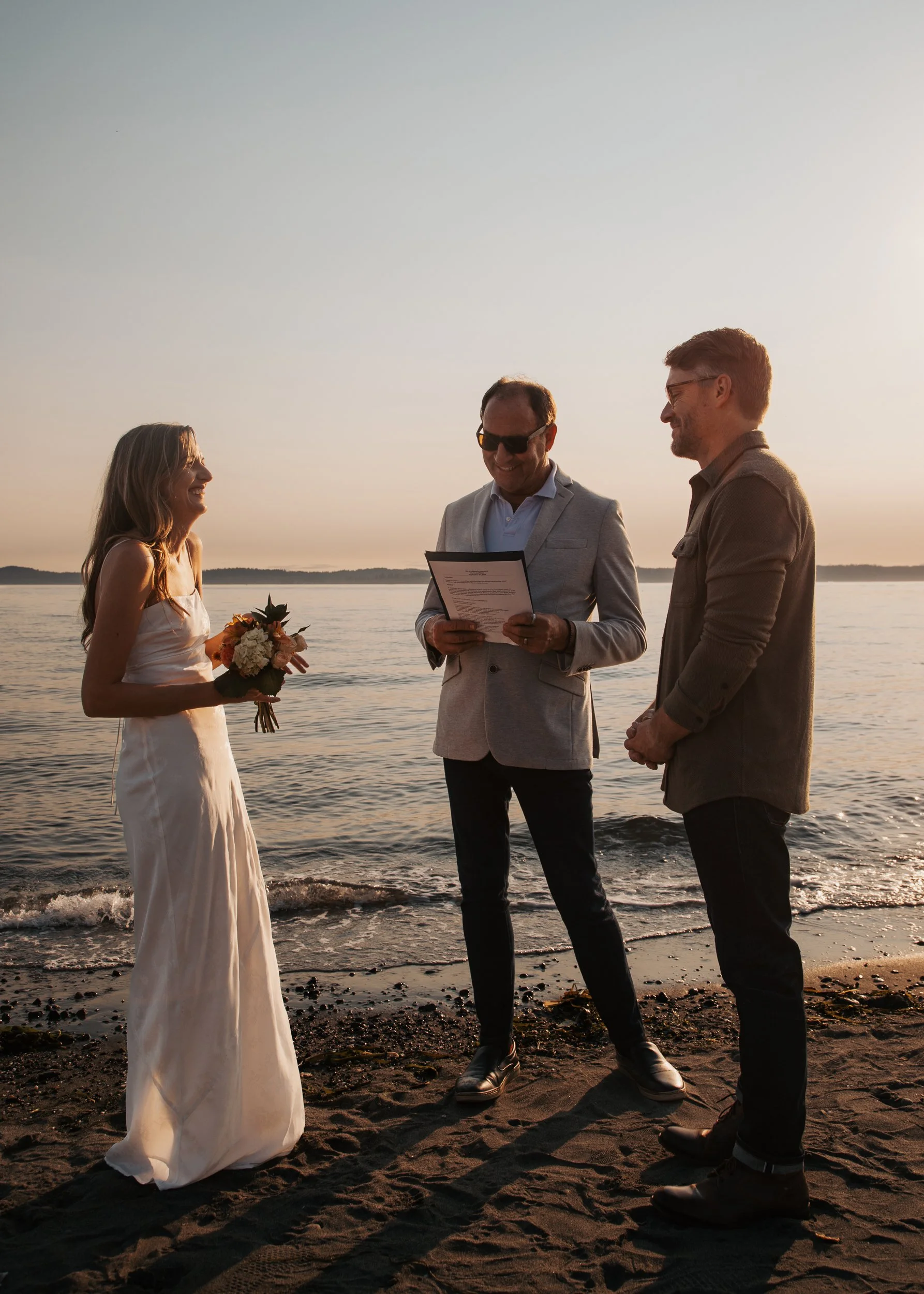 A wedding ceremony on the beach at sunset with a woman in a white dress holding a bouquet. Discovery Park Beach Lighthouse elopement