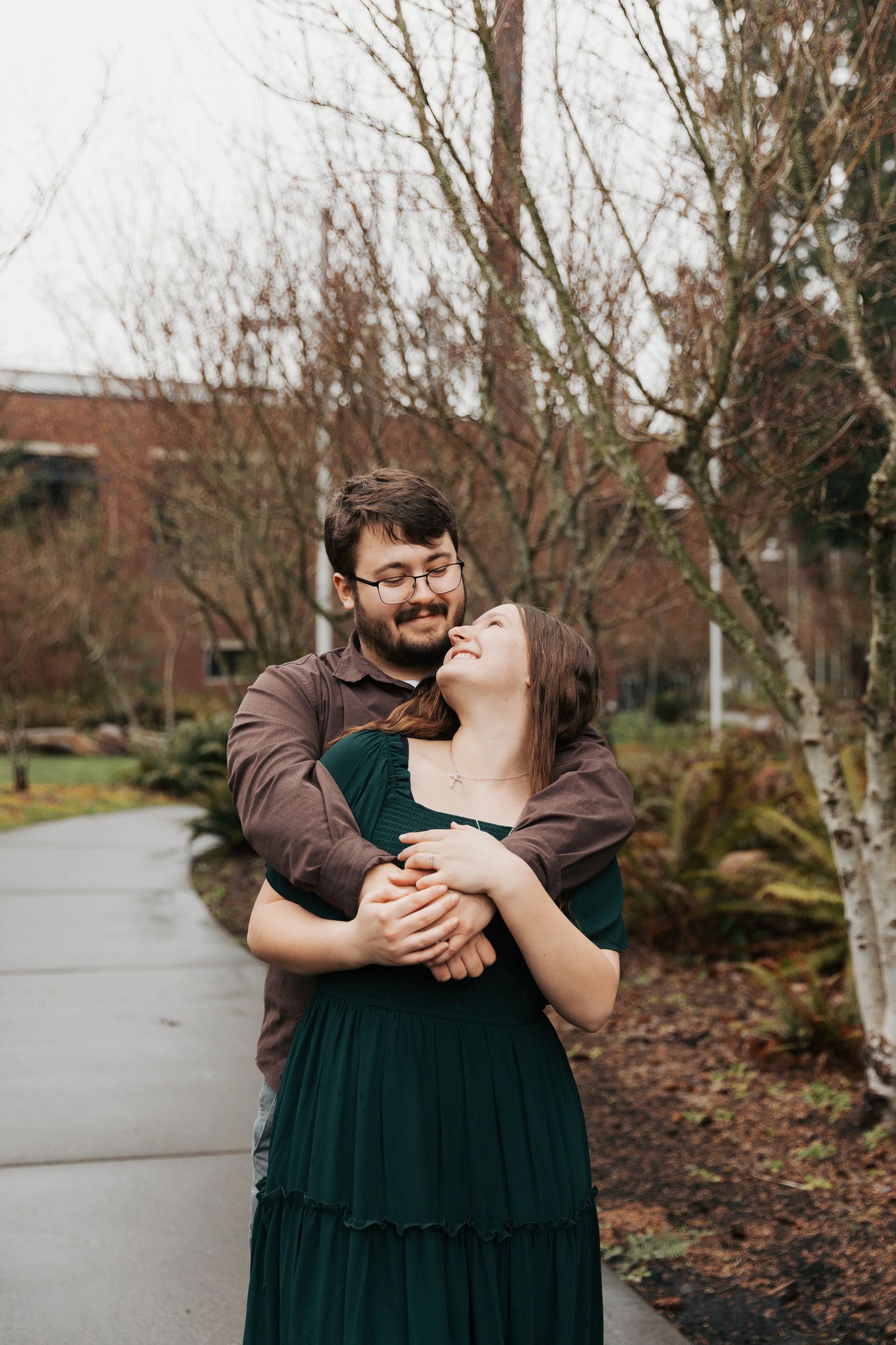 A man and woman embracing outdoors on a cloudy day, with leafless trees and a brick building in the background.