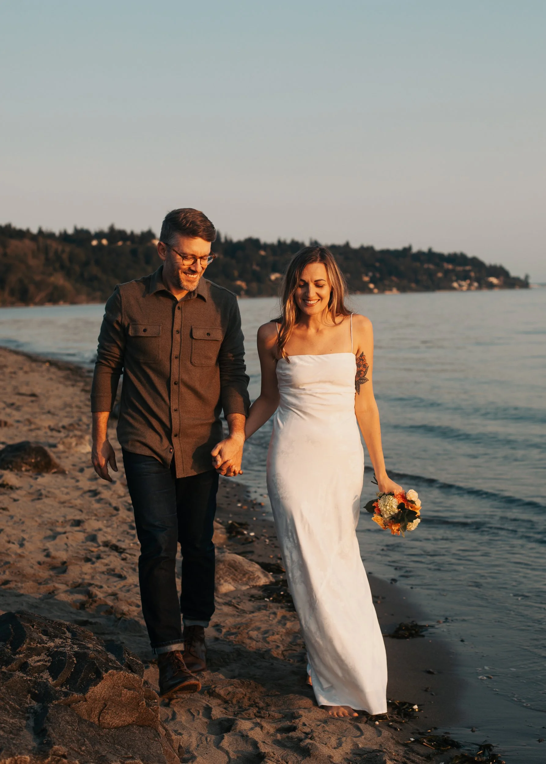 A couple holding hands walking on a beach at sunset, with the woman in a white dress holding a bouquet of flowers. Discovery Park Beach Lighthouse elopement